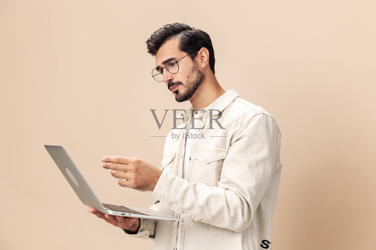 一位时尚男士,戴着眼镜,手持笔记本电脑,神情沉思,穿着白色T恤,背景为米色,展现出时尚的穿衣风格,空间感十足。照片摄影图片