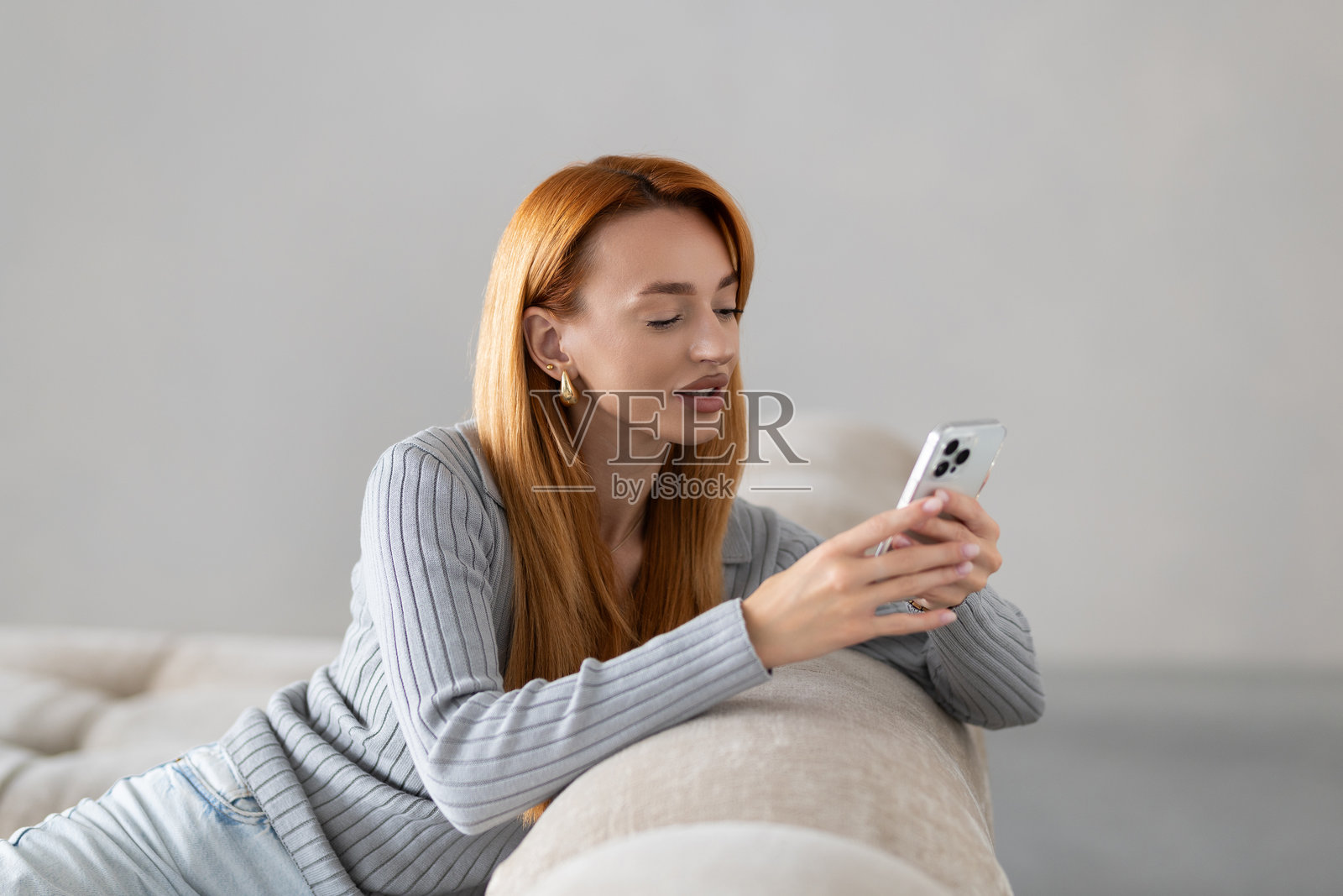 在家客厅的沙发上躺着一位快乐的女性,正在使用智能手机。照片摄影图片