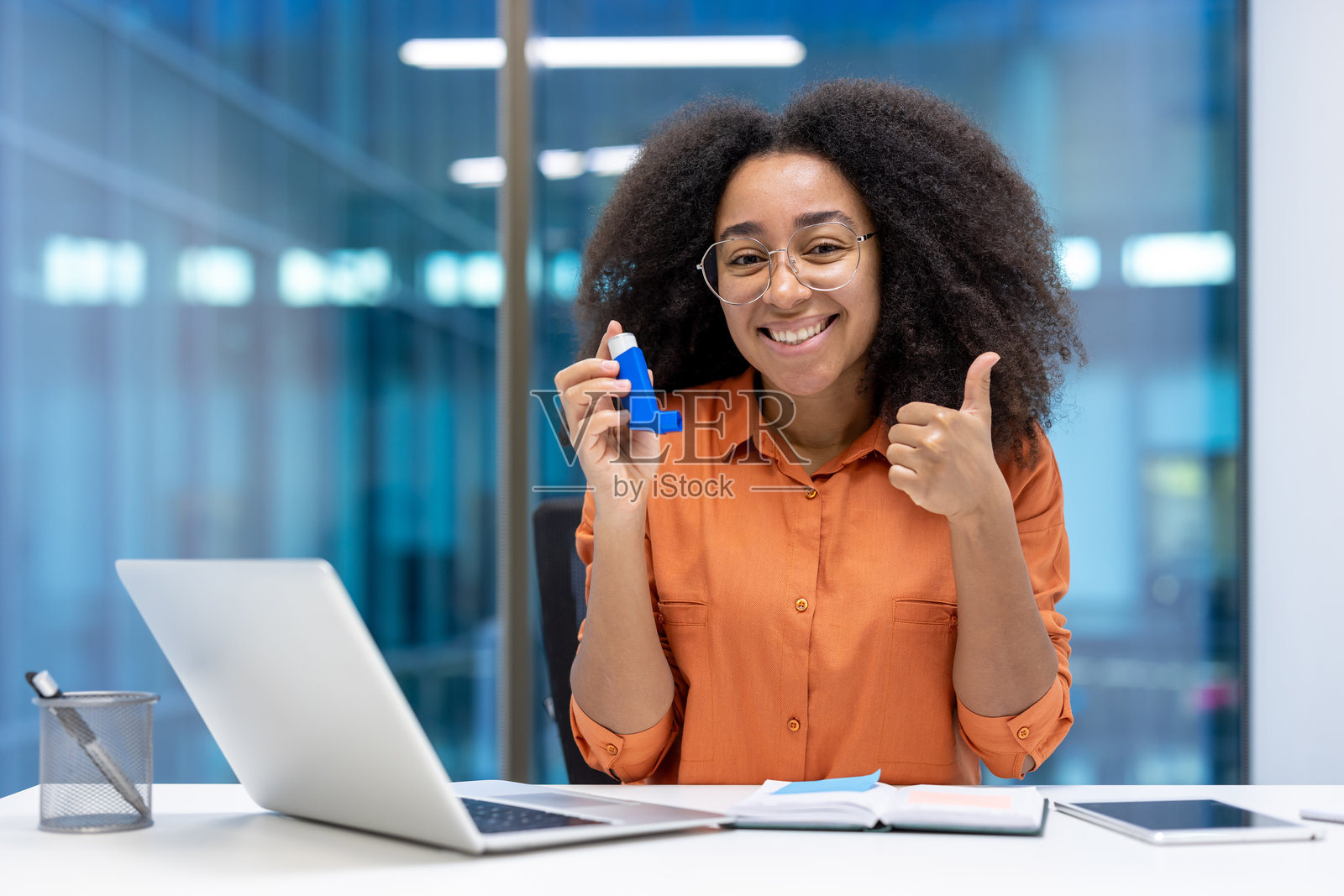 微笑的女性在办公室环境中自信地握着吸入器,竖起大拇指,倡导健康意识和积极态度,营造多元化的工作场所氛围。照片摄影图片
