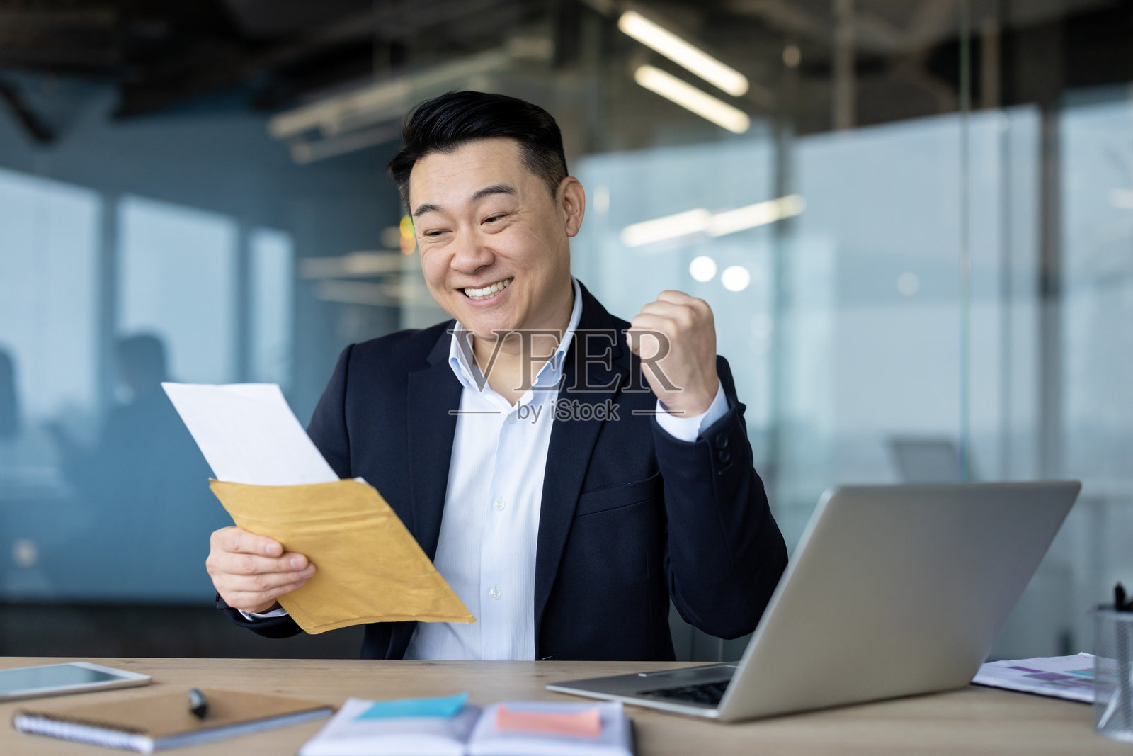 亚洲年轻男性商人在办公室的桌子旁坐着,手中拿着一个打开的信封,正在阅读一封提醒信,因收到好消息而感到高兴,做出胜利的手势。照片摄影图片