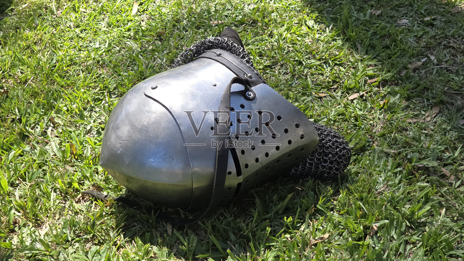 中世纪骑士头盔和链甲 lying on grass. 头盔顶部圆润,面罩有孔,配有带子固定。照片摄影图片