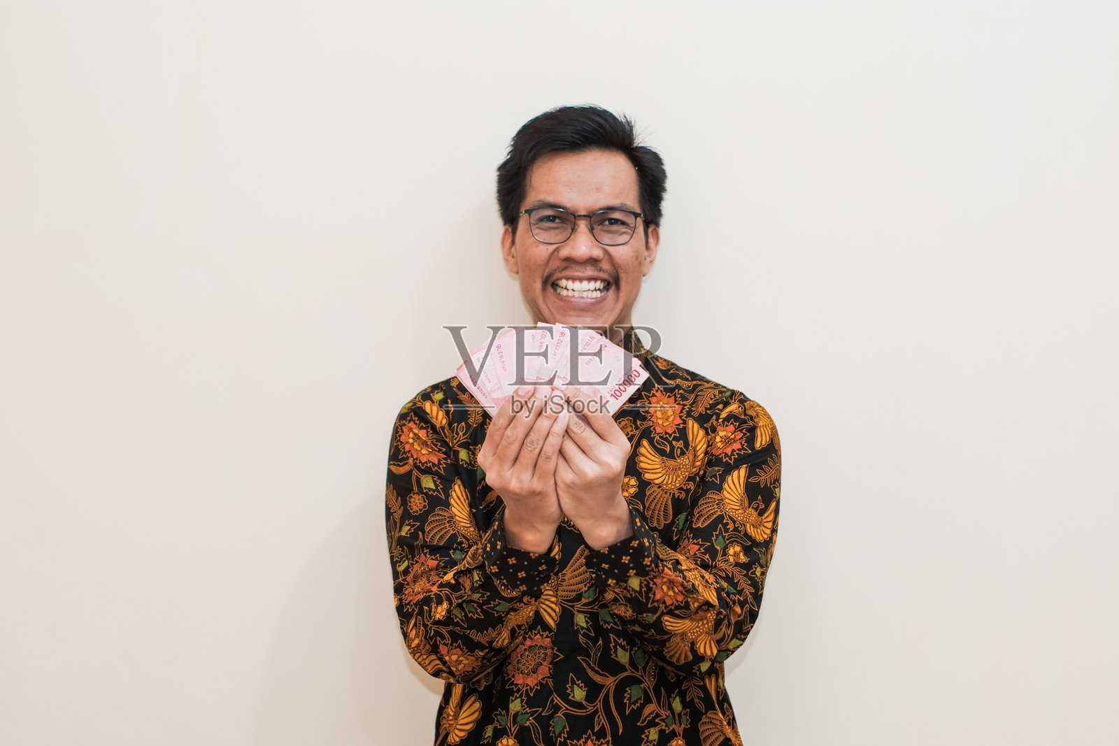 一位快乐微笑的东南亚男子,穿着巴迪克衬衫,戴着眼镜,手里拿着印尼卢比现金。照片摄影图片