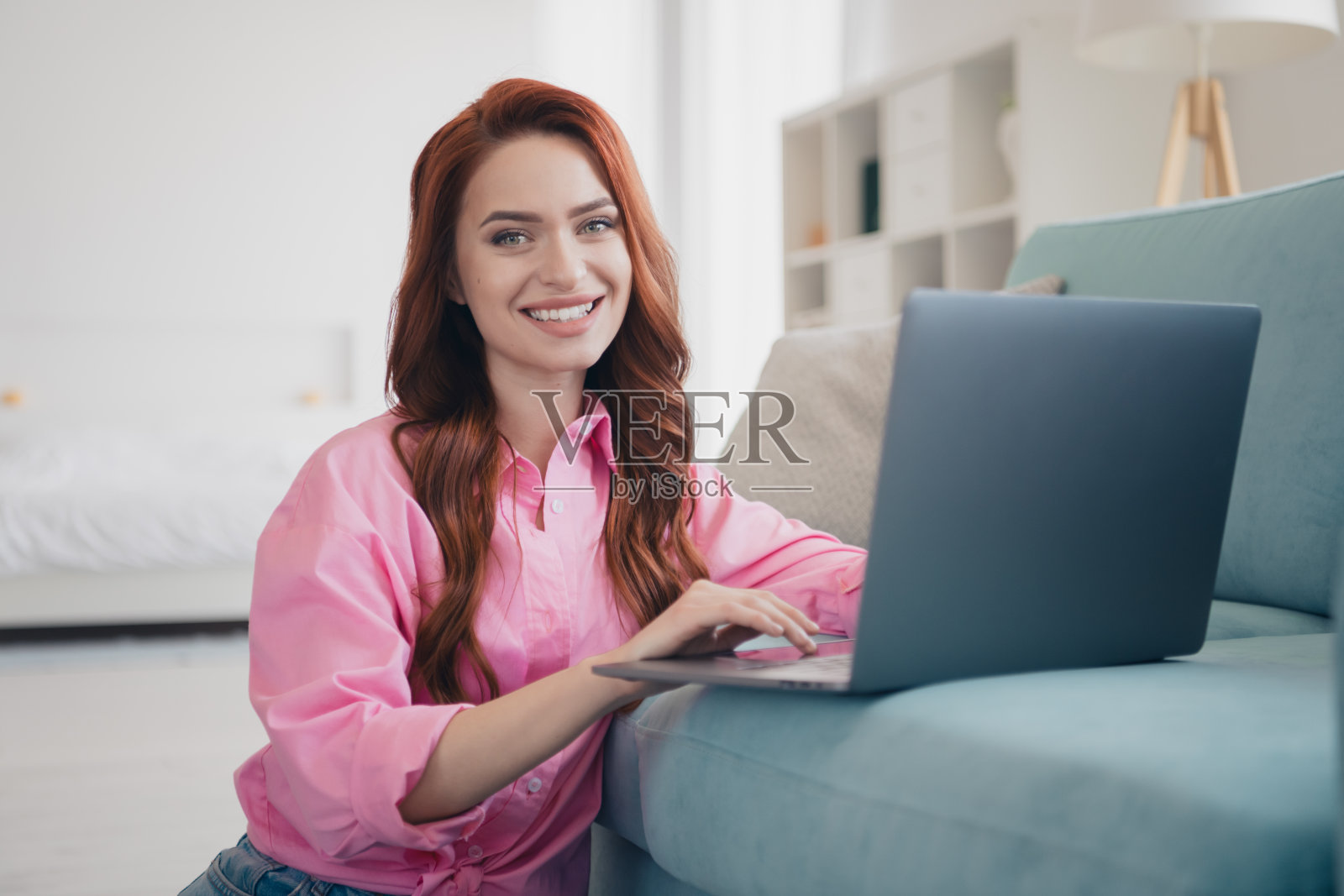 年轻漂亮的女性露齿而笑,穿着粉色衬衫,使用笔记本电脑,坐在现代明亮的公寓室内。照片摄影图片