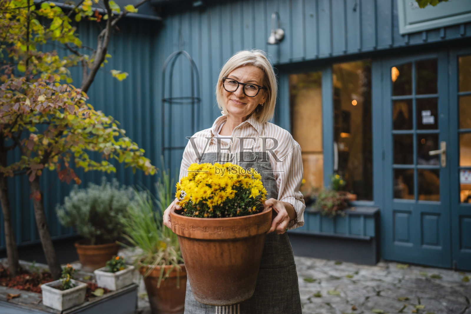 成熟的女服务员在乡村商店外拿着一盆黄色花卉照片摄影图片