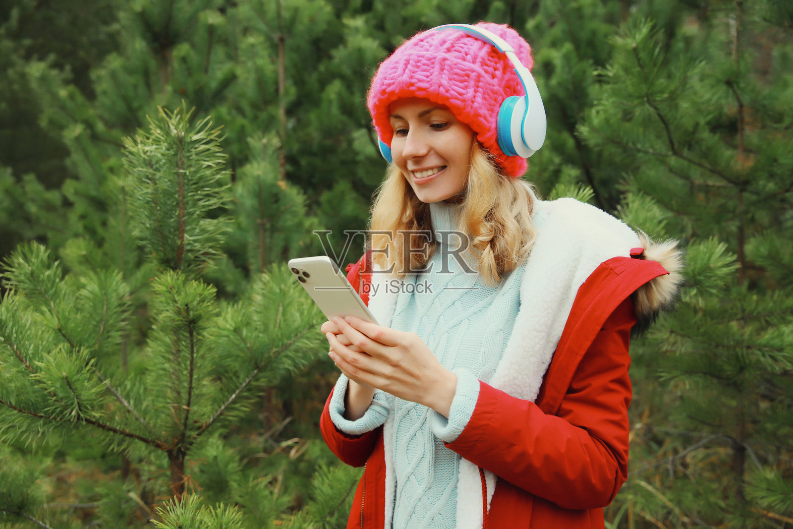 快乐的年轻女性在冬季森林中戴着耳机用手机听音乐,身旁有一棵圣诞树。照片摄影图片