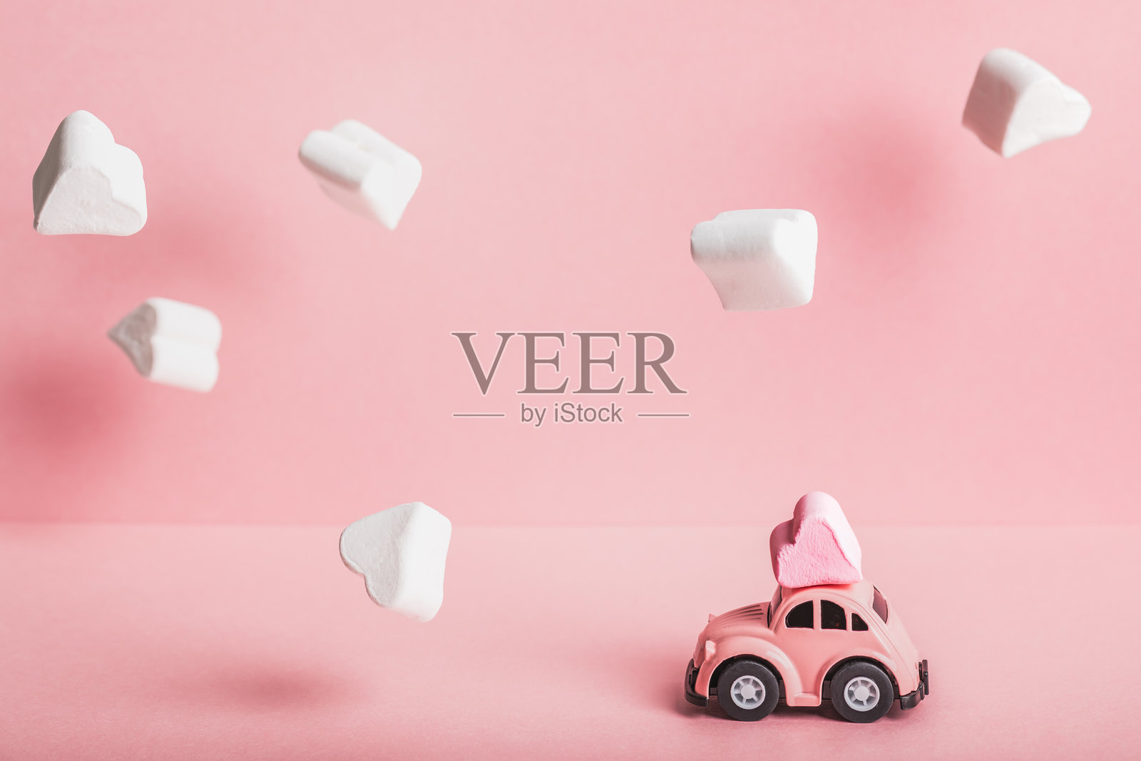 一辆小巧的粉色汽车模型在粉色背景上载着心形棉花糖,周围是棉花糖云朵。适合情人节、生日、婚礼日和母亲节的概念。照片摄影图片