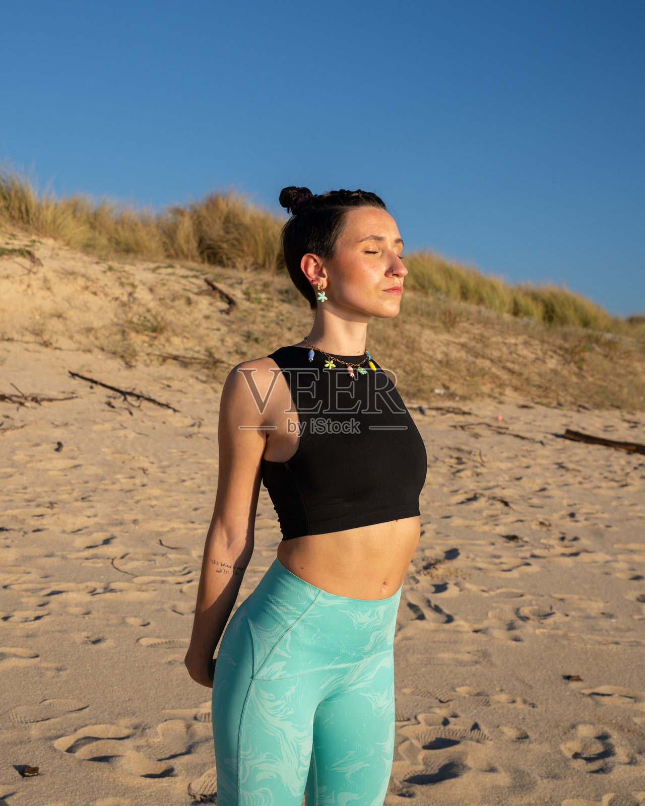 穿着运动服和五彩耳环的女性在阳光明媚的海滩上闭眼练习瑜伽,背景是沙子和沙丘。照片摄影图片