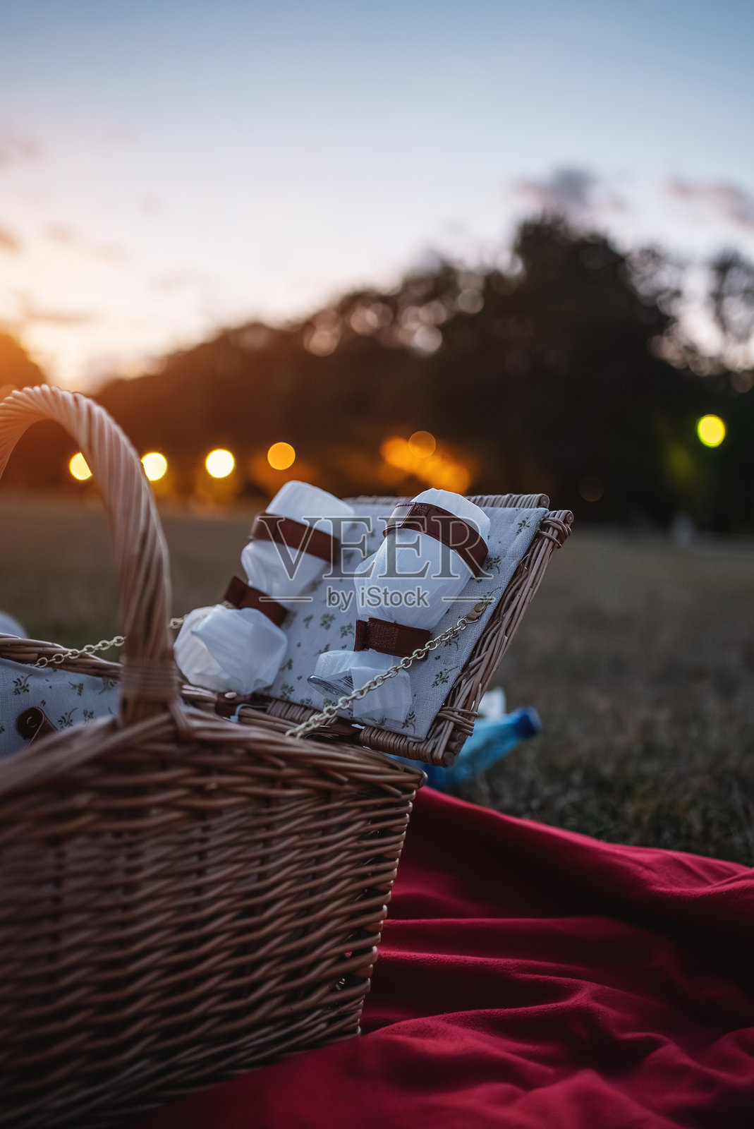 温馨的野餐篮里装满了白色杯子、酒杯和小吃,放在公园里鲜艳的红色毯子上,夕阳下宁静的氛围非常适合享受户外的夜晚。照片摄影图片