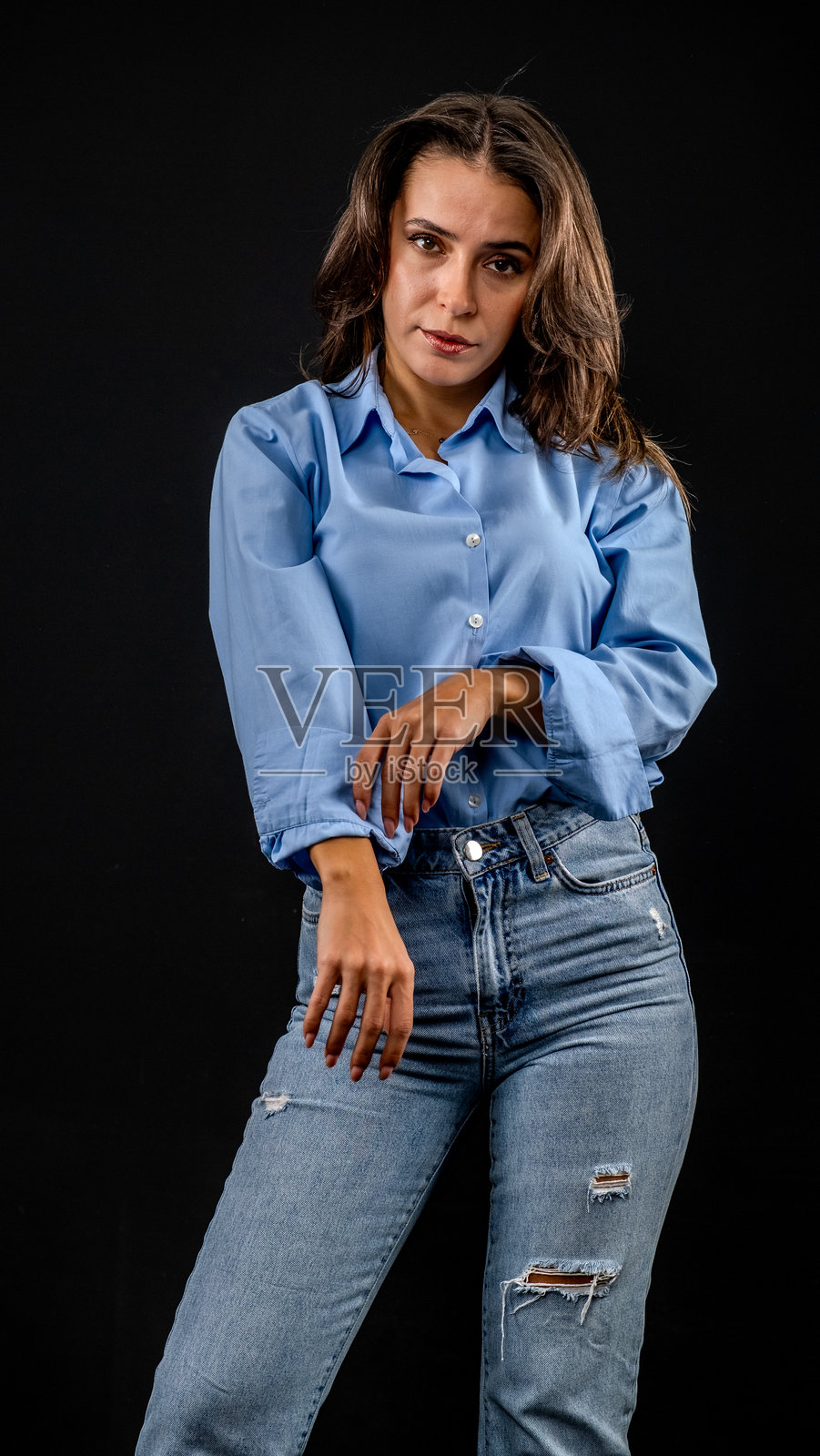 在黑色背景的摄影棚中,一位年轻的棕发女性穿着蓝色衬衫和牛仔裤,以艺术风格摆姿势。照片摄影图片