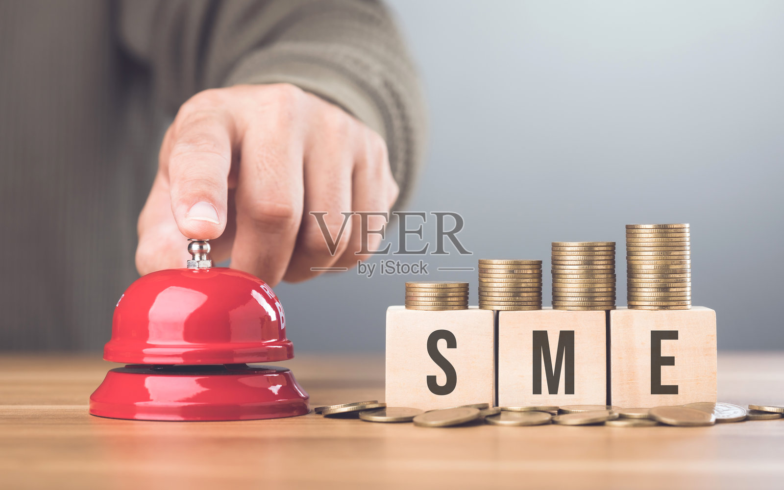 一只手按下一个红色铃铛,旁边是一堆硬币和一个标有“SME”的木块,象征着新小企业冒险的开始以及早期财务规划的重要性。照片摄影图片