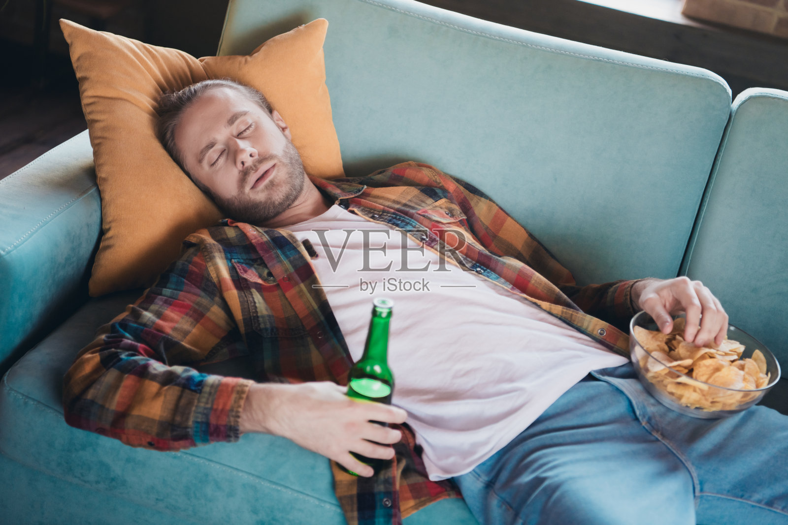 放松的年轻男子在阳光明媚的客厅沙发上打盹,穿着格子衬衫,享受宁静的周末下午。照片摄影图片