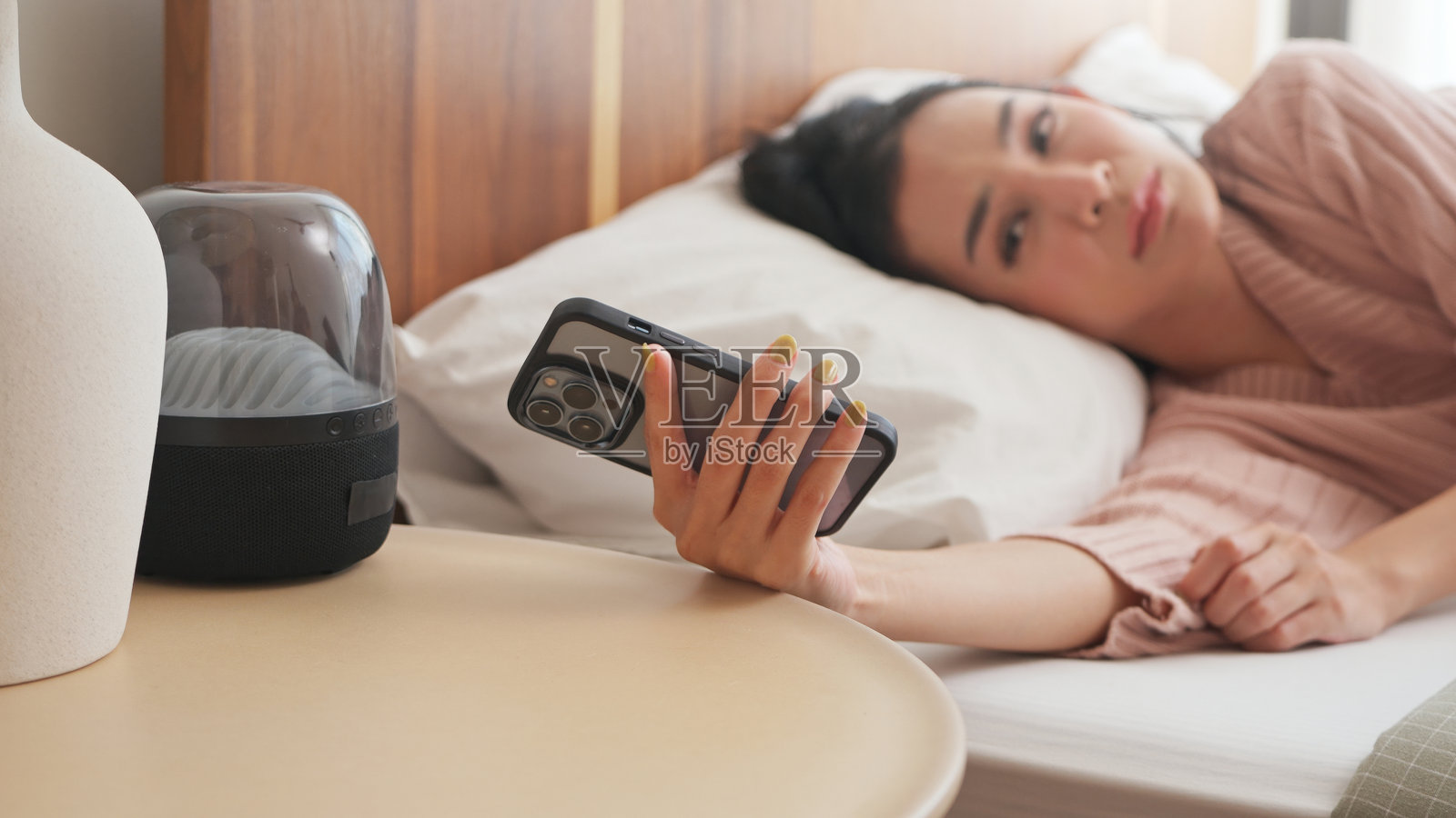 困倦的年轻亚洲女性躺在床上,手握智能手机,神情沉思,早晨的卧室。照片摄影图片