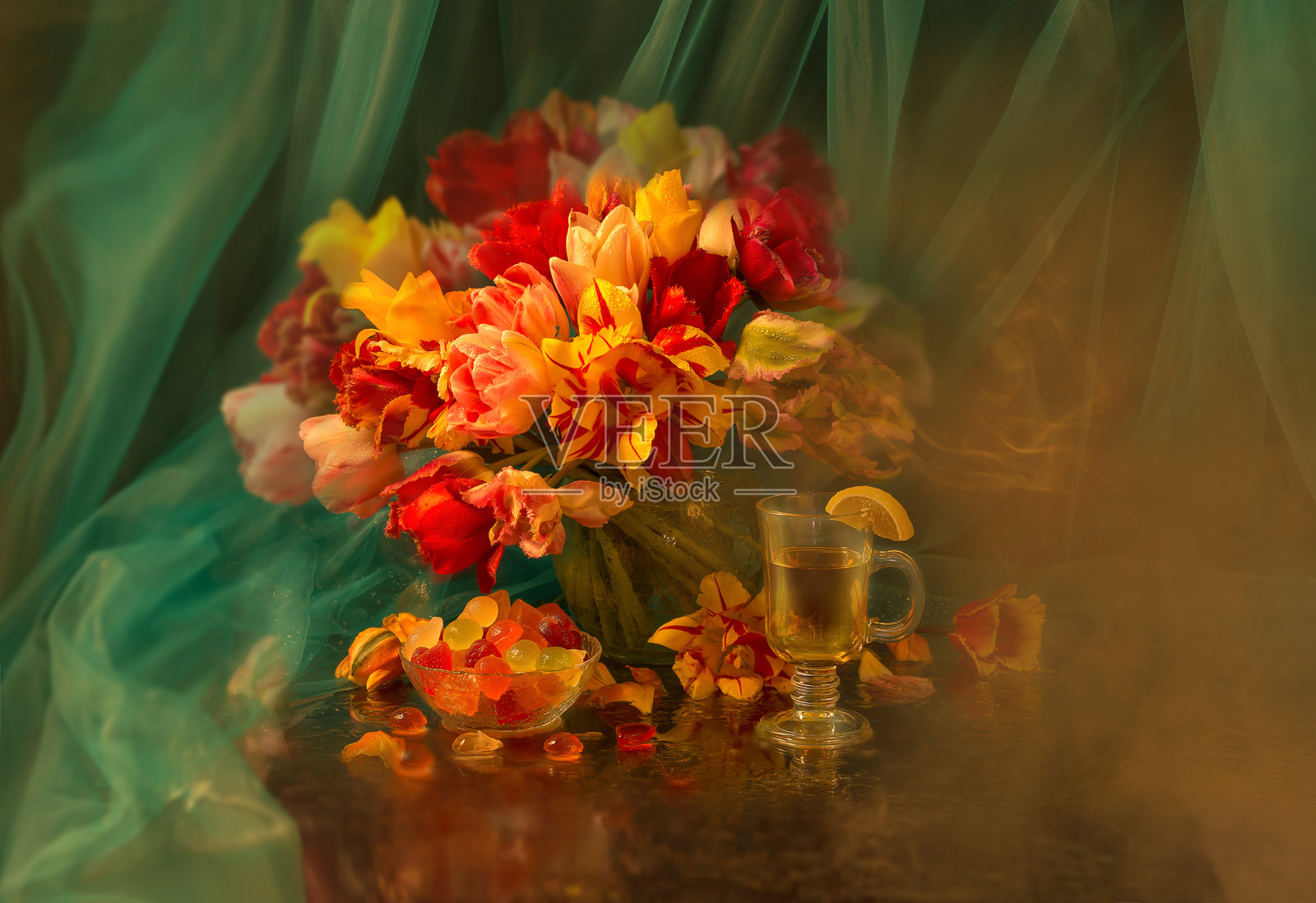 静物画:一束鲜艳美丽的郁金香放在透明花瓶中,背景为绿色。旁边有一杯茶和五彩缤纷的果冻。照片摄影图片