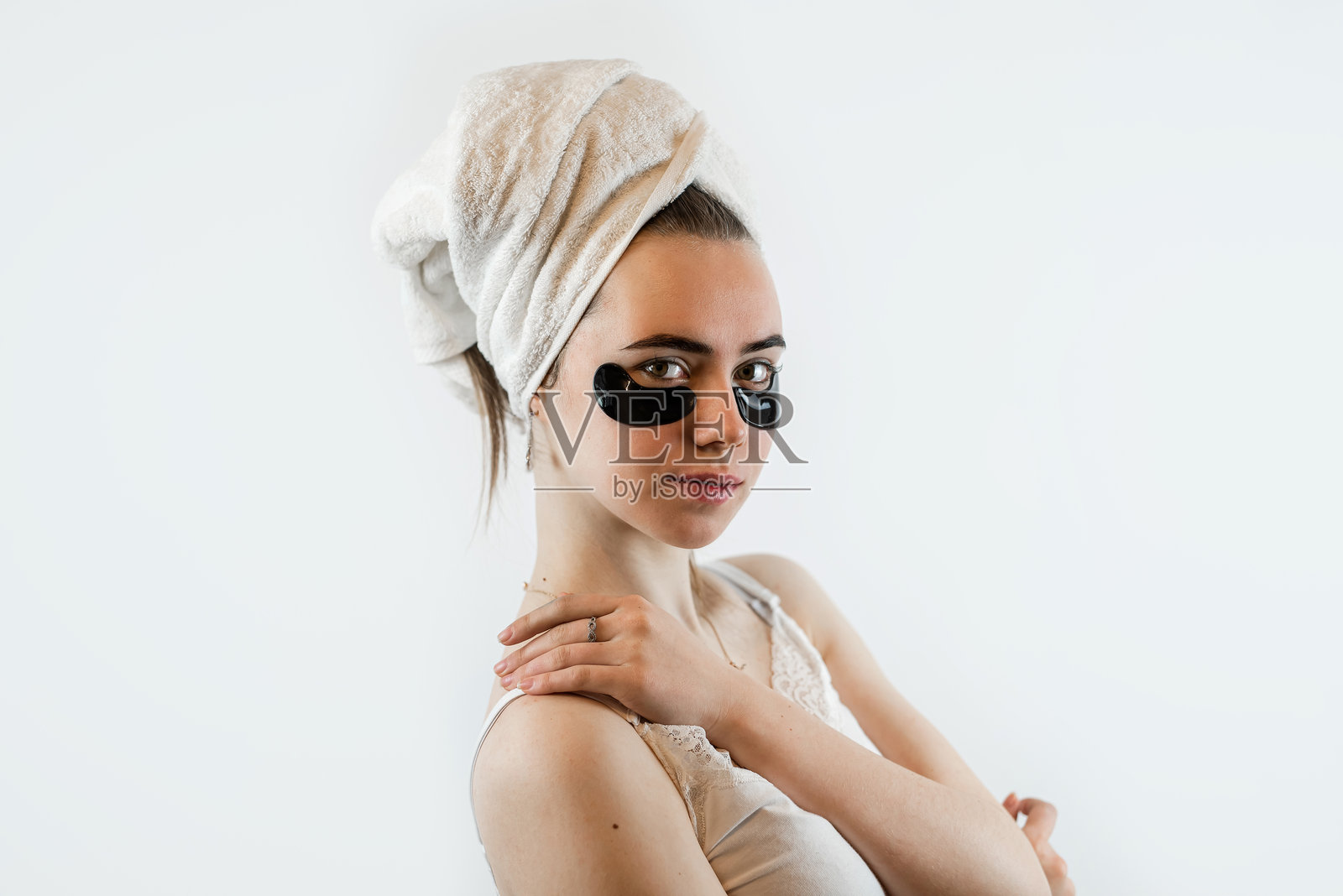 年轻漂亮的女士用白色毛巾包裹着身体,在眼下涂抹胶原蛋白水凝胶贴片,以平滑肌肤,背景为白色。照片摄影图片