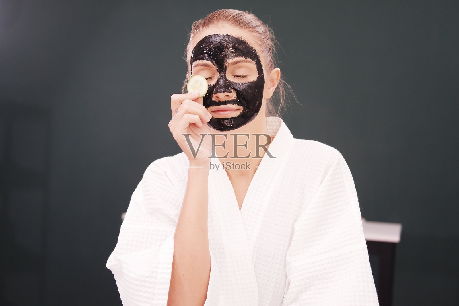 微笑的白人少女穿着浴袍,手持切片黄瓜和黑色粘土面膜,放松地进行面部护理,享受水疗治疗。照片摄影图片
