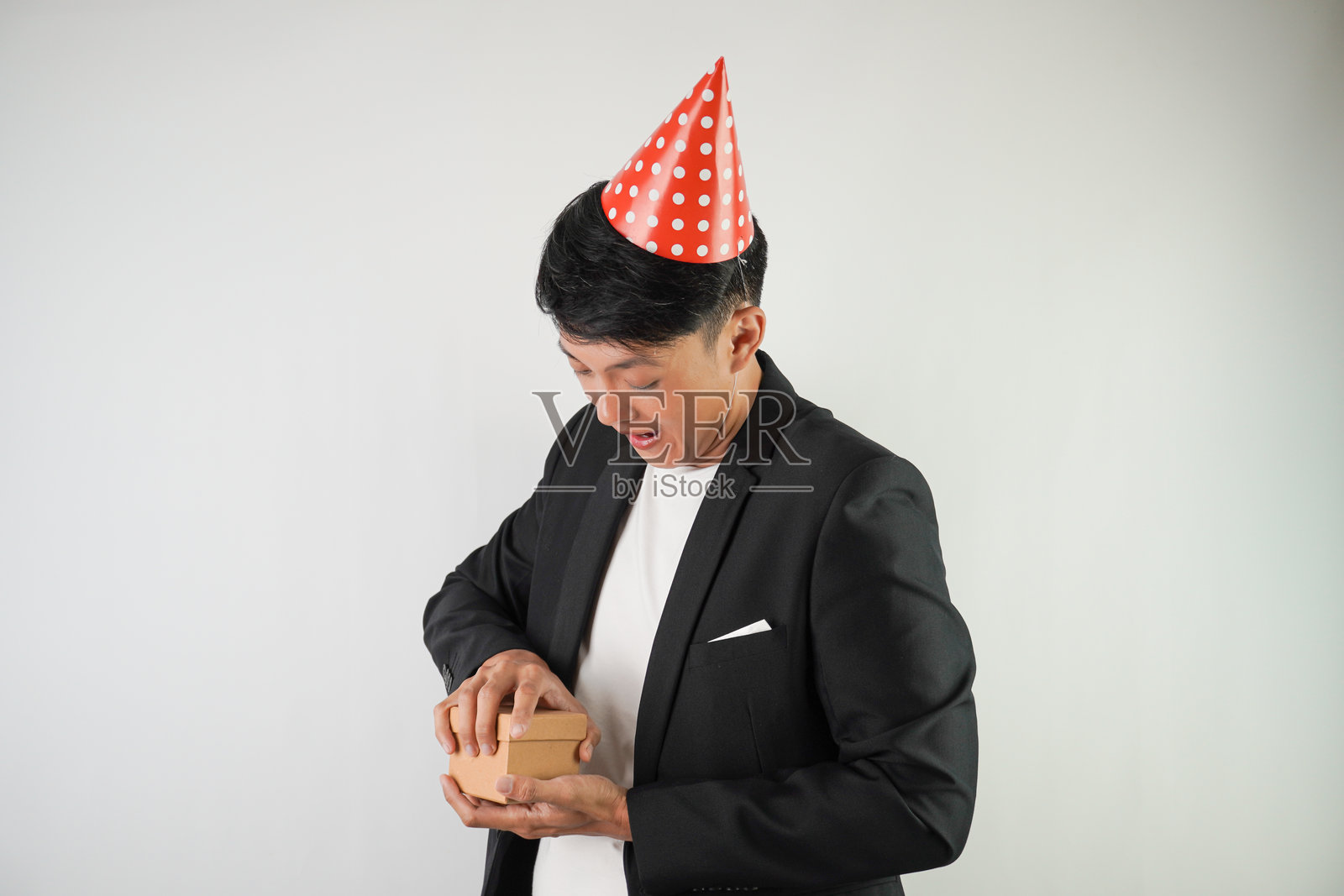 兴奋的年轻亚洲男子戴着红色派对帽,穿着白衬衫和黑色西装,正在打开一个小礼品盒,背景为白色。适用于圣诞节和新年派对的概念。照片摄影图片