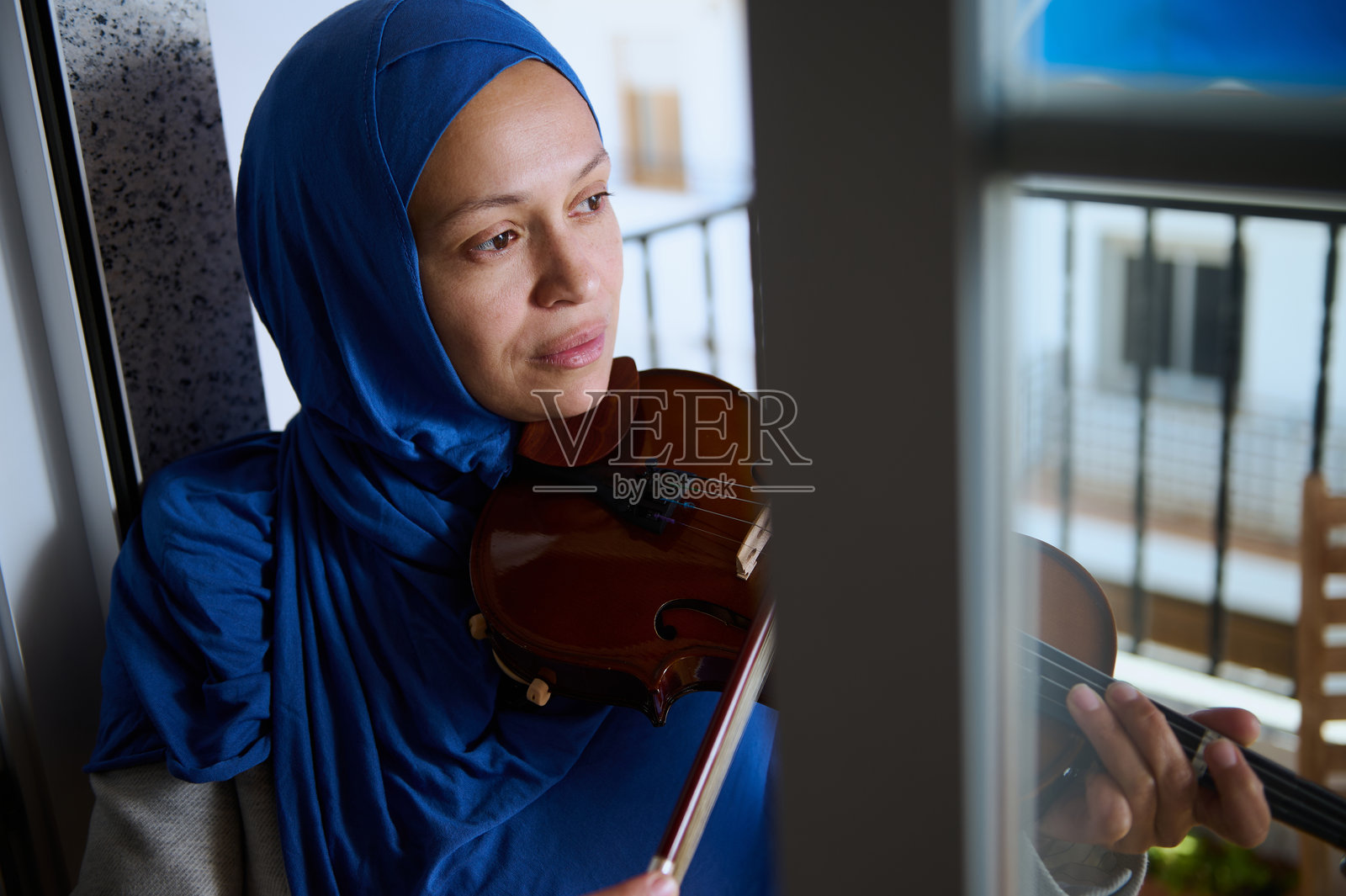 身穿蓝色头巾的女性在窗边沉思地拉小提琴照片摄影图片