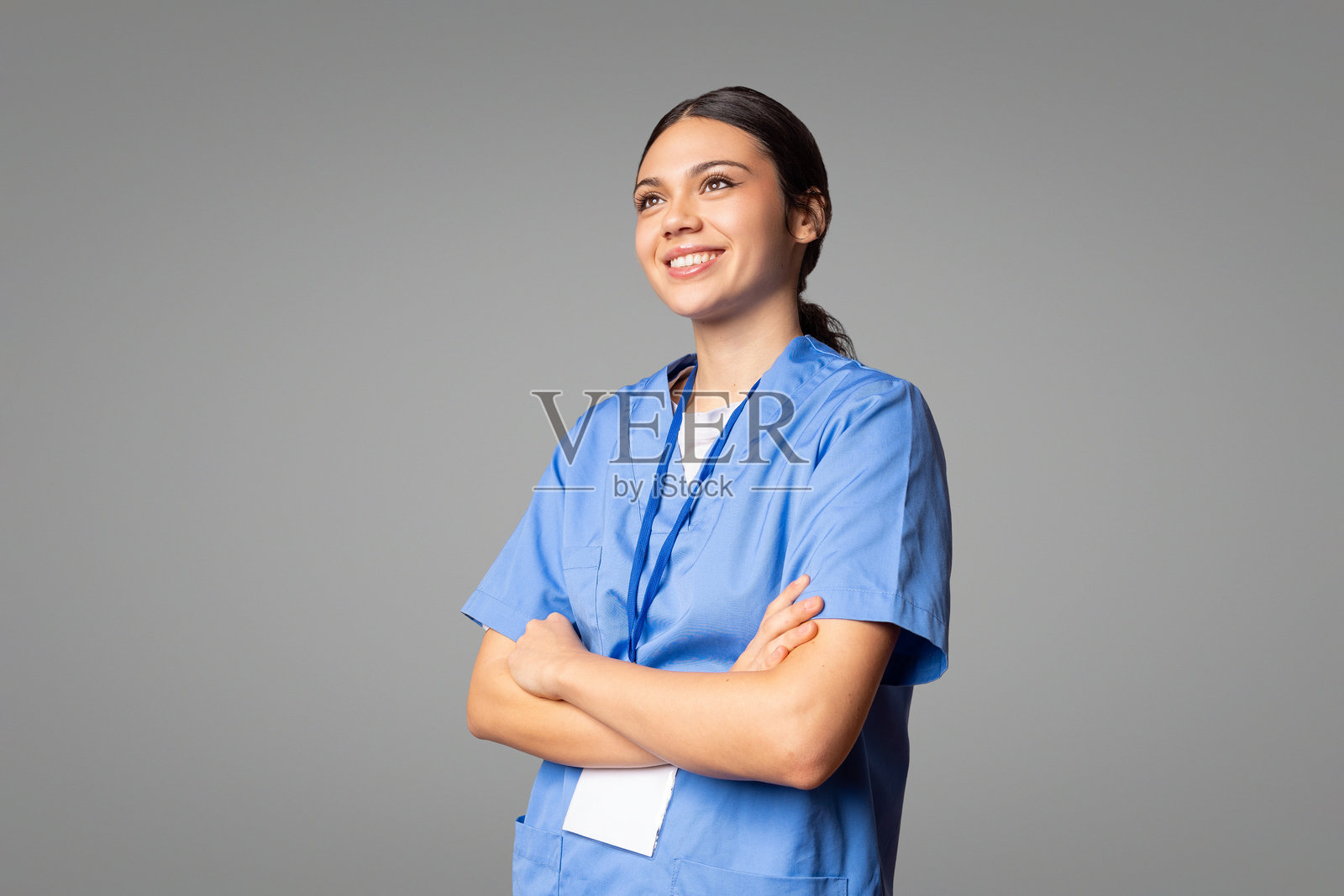 微笑着的年轻女医生,双臂交叉,仰望着,穿着蓝色制服和徽章。照片摄影图片