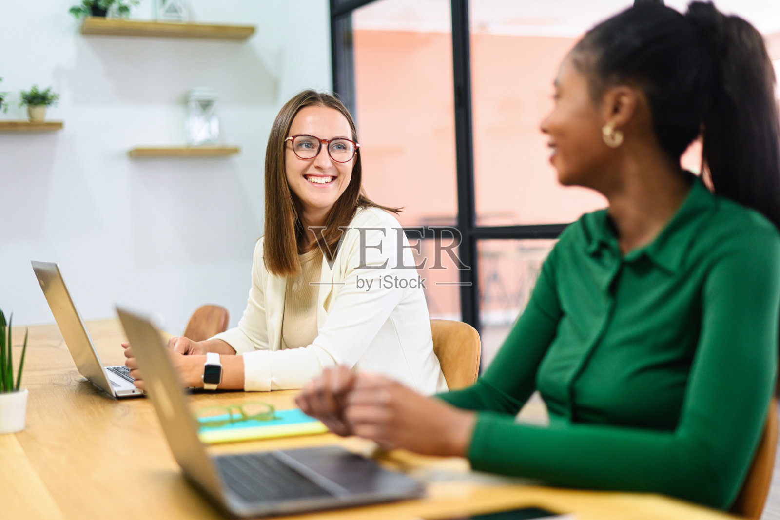 两位不同背景的女性员工穿着休闲商务服装,在现代会议室中一起使用笔记本电脑,友好地分享计划和想法,营造出良好的工作氛围。照片摄影图片