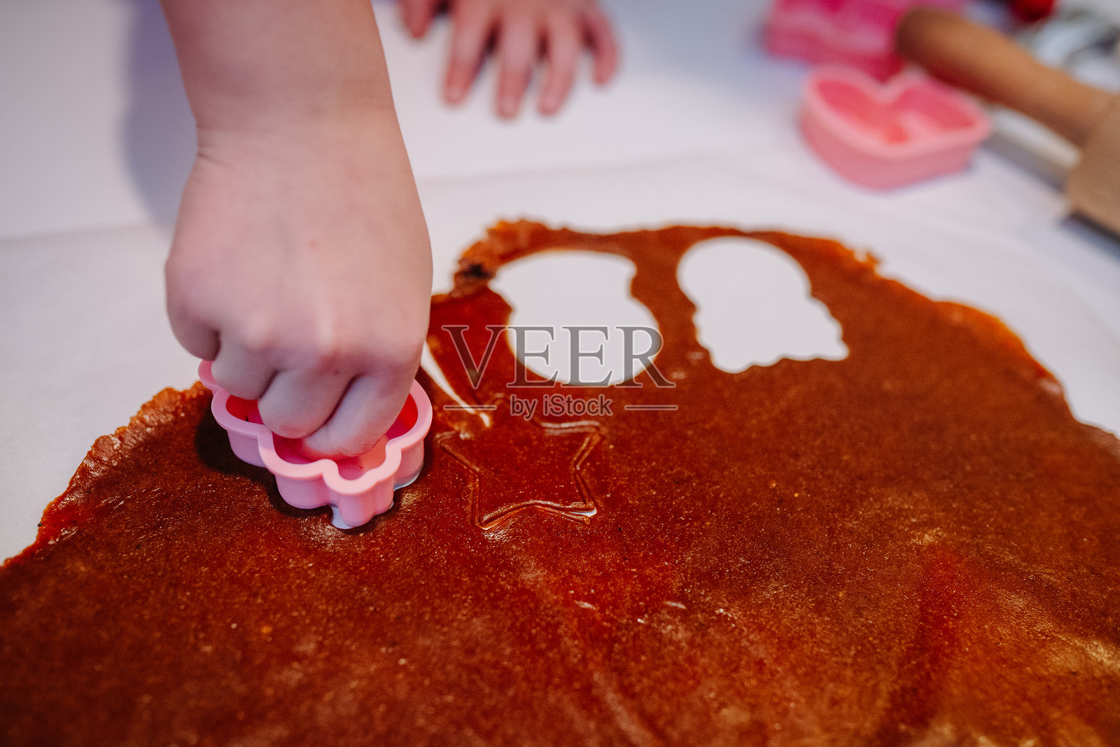 孩子的手在擀平的棕色面团上使用粉色饼干模具,正在烘焙纸上制作节日形状。照片摄影图片