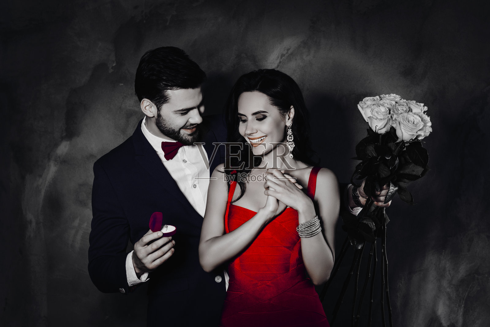 时尚绅士蓄着胡茬,向兴奋快乐的女士赠送一枚镶有钻石的金戒指,女士手中捧着白玫瑰,情侣在灰色背景下独立成图。照片摄影图片