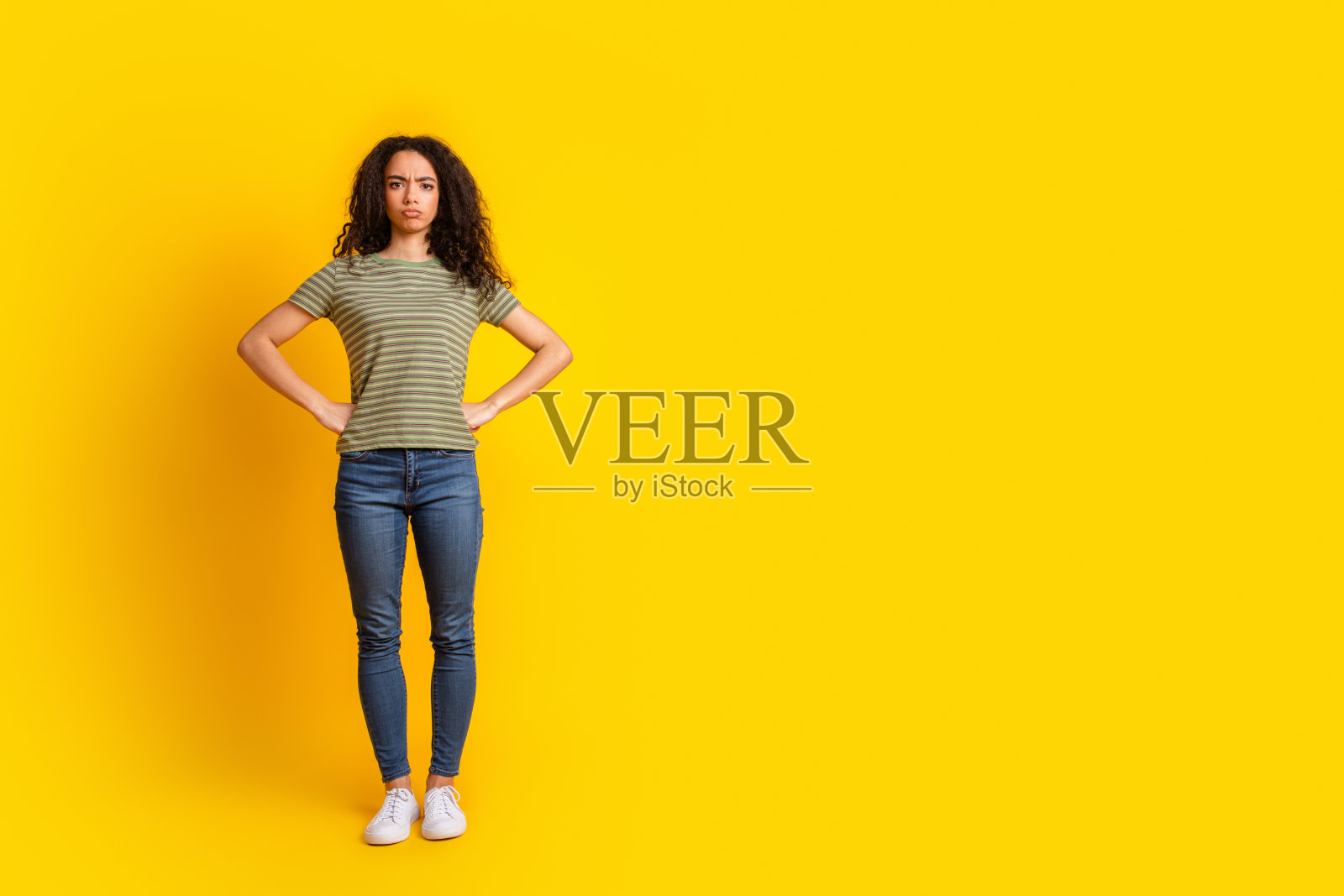 全身照片,严肃自信的女孩穿着条纹T恤,双手叉腰,背景为鲜艳的黄色。照片摄影图片