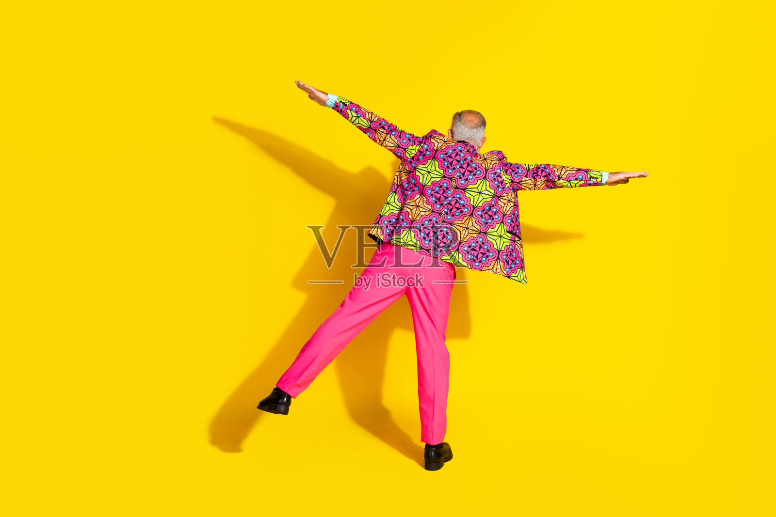 穿着粉色裤子和多彩西装的时尚老年男子单腿站立,背面全身肖像,背景为黄色。照片摄影图片