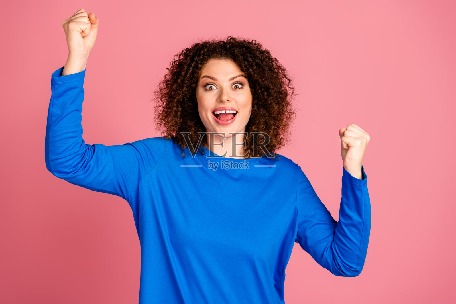 兴奋的年轻女性,卷发,穿着蓝色毛衣,在粉色背景前庆祝照片摄影图片