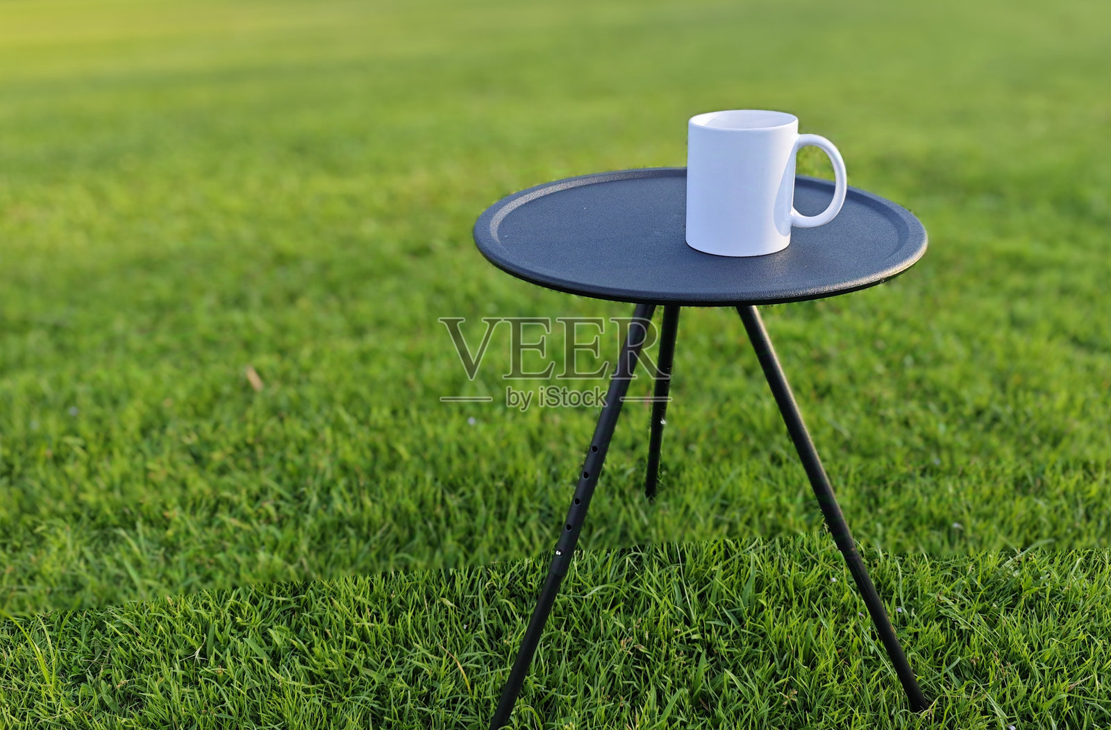 两只白色杯子放在黑色圆桌上,桌子下是绿色的草地。照片摄影图片