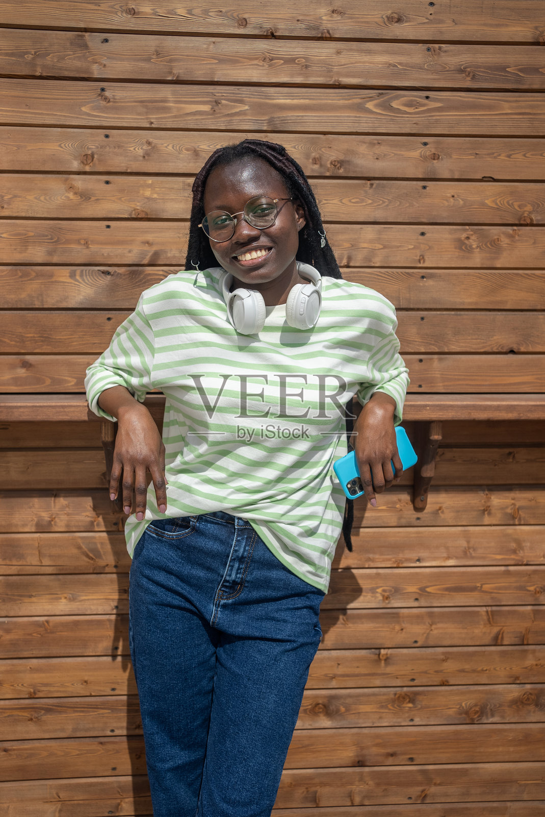 微笑的黑人女性戴着耳机在户外放松,手持智能手机靠在木质背景上照片摄影图片