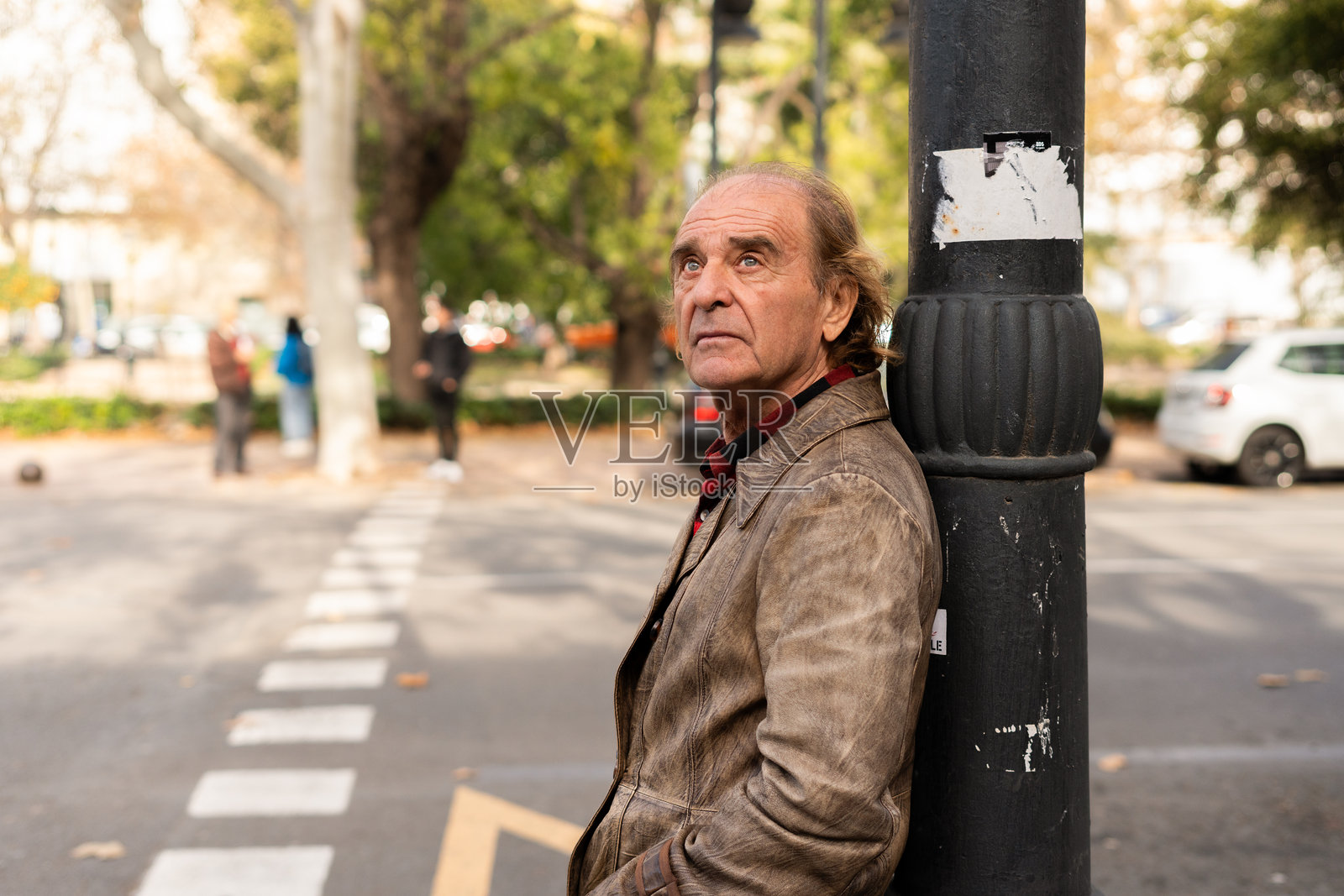 一位老年男子靠在城市街道的柱子上,目光投向远方。照片摄影图片