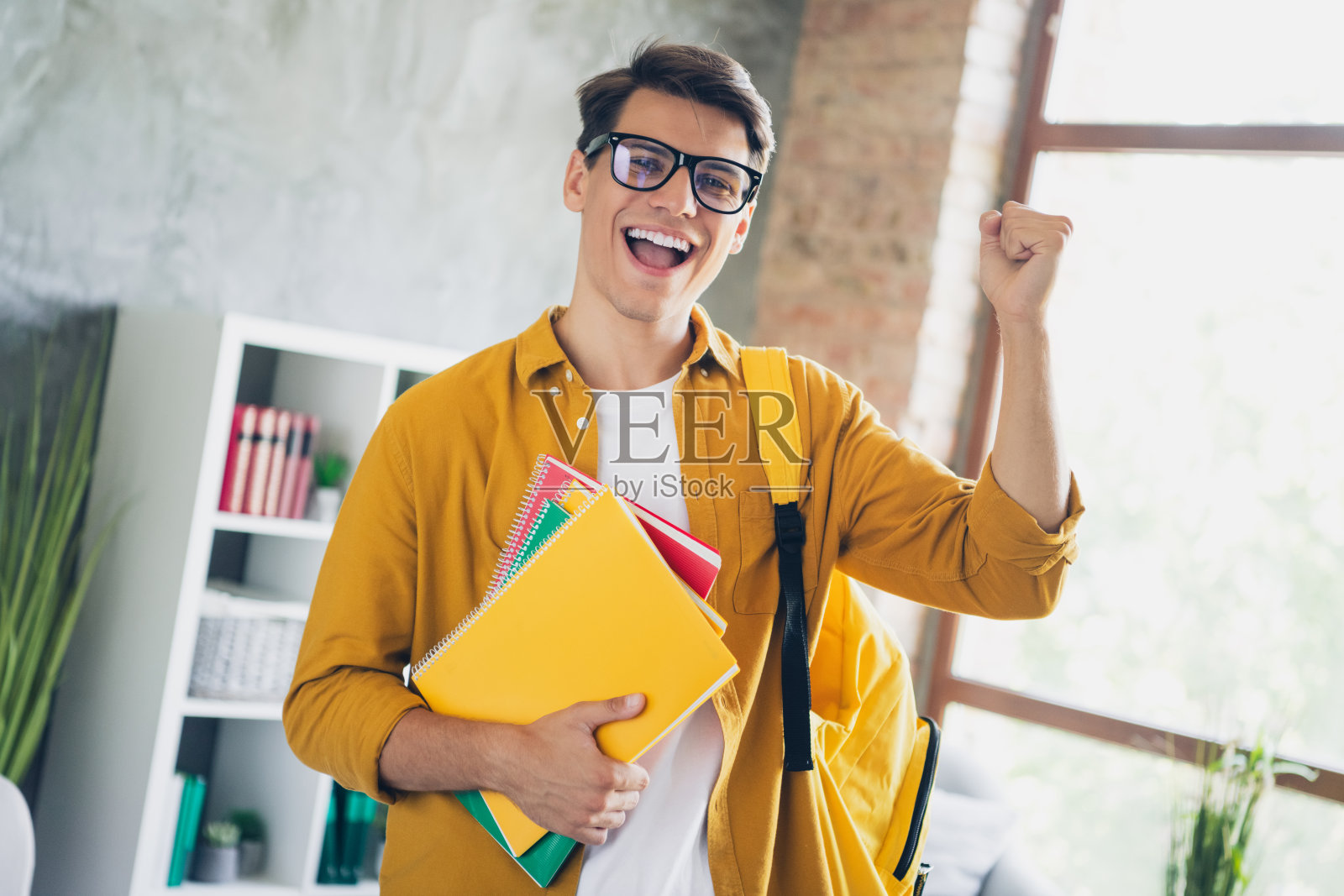 快乐的年轻男子戴着眼镜,穿着黄色衬衫,在日光下的家居环境中手持笔记本。照片摄影图片