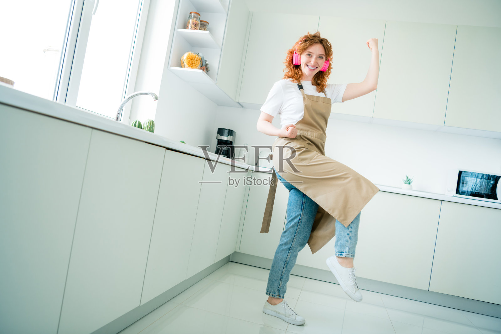 快乐的年轻女性,红发,穿着休闲风格的围裙,在现代厨房中跳舞,感到愉快和充满活力。照片摄影图片