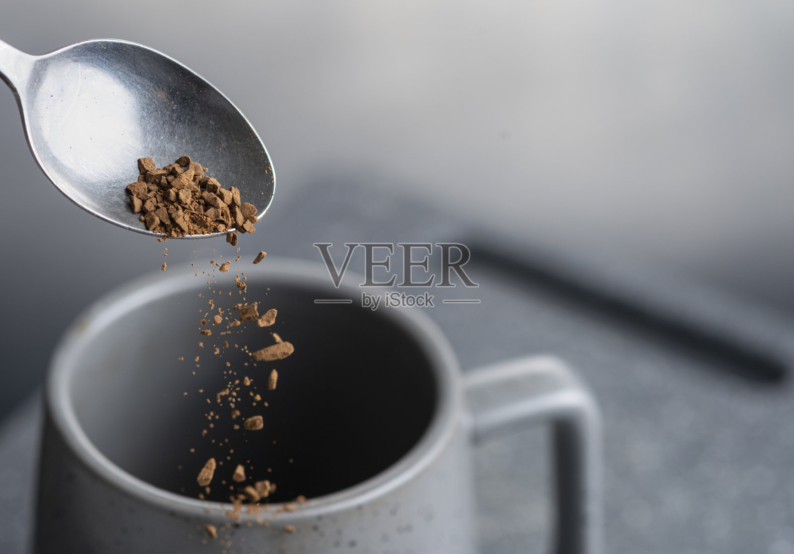 从银色茶匙上掉落的冻干速溶咖啡颗粒,落入空杯中,背景为石板灰色。照片摄影图片