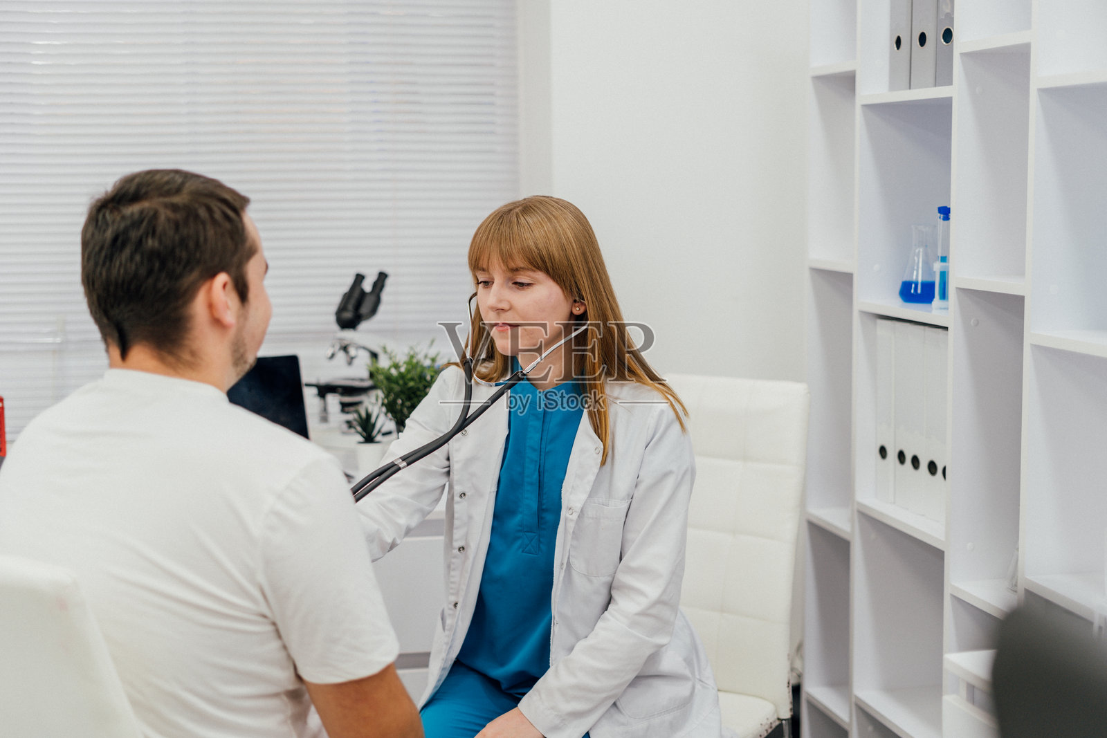 一位佩戴听诊器的女医生在临床咨询中检查一名男性患者。明亮整洁的医疗办公室突显了专业性和对患者的关怀。照片摄影图片