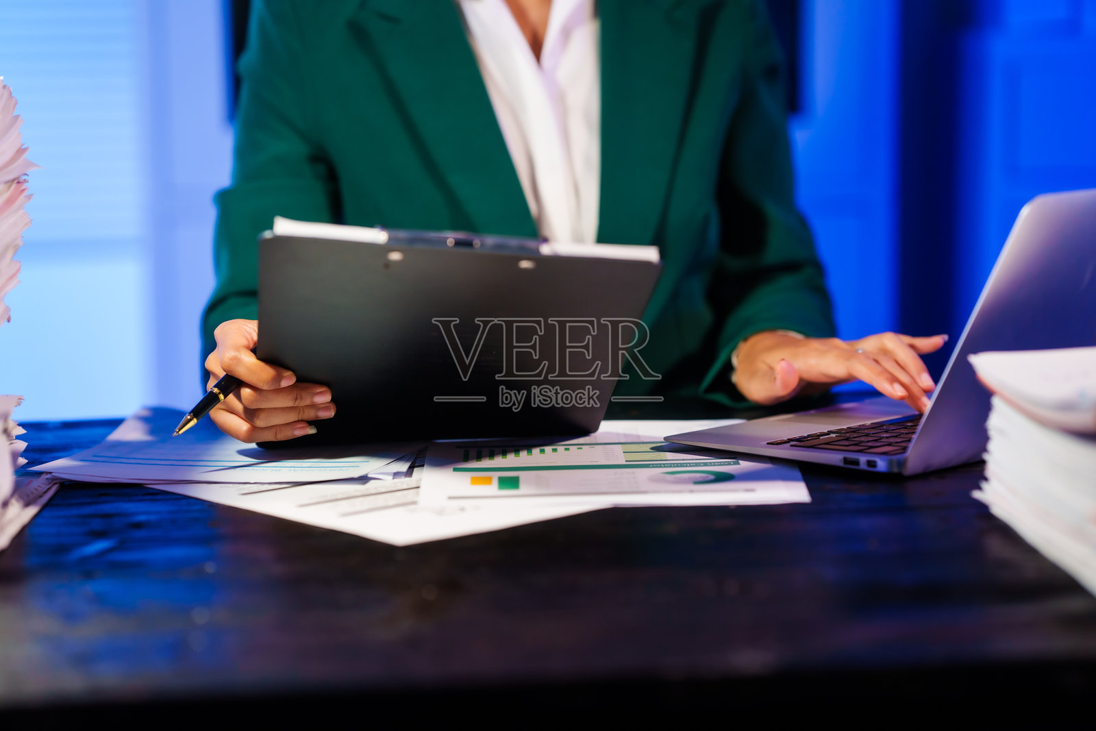 一位亚洲女商人在夜晚加班,坐在办公桌前,通过电脑或笔记本电脑查阅大量文件。面对办公室问题,她一边处理繁重的工作,一边在线聊天。照片摄影图片