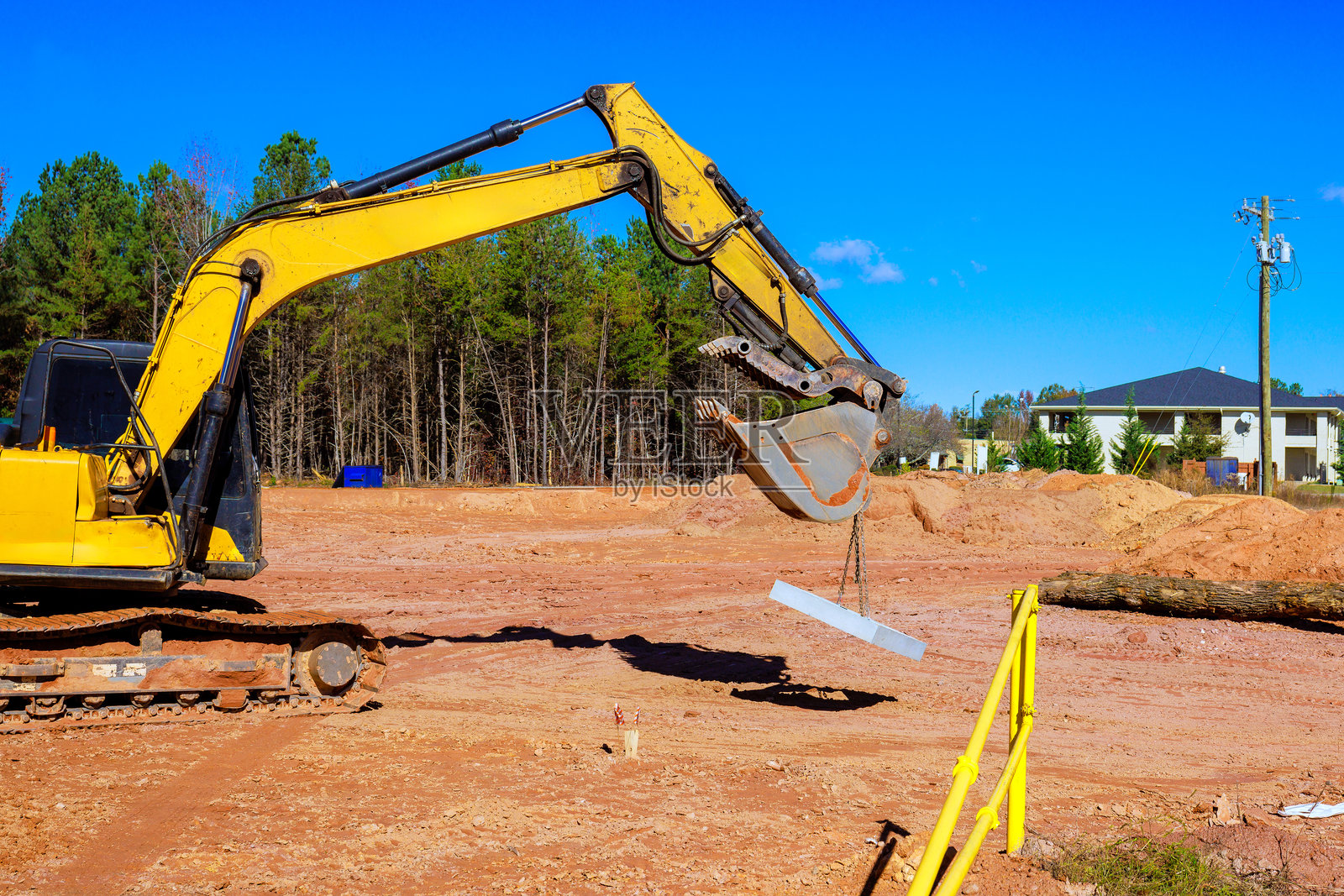 挖掘机在施工现场抬起一根梁,四周环绕着树木,蓝天晴朗。照片摄影图片