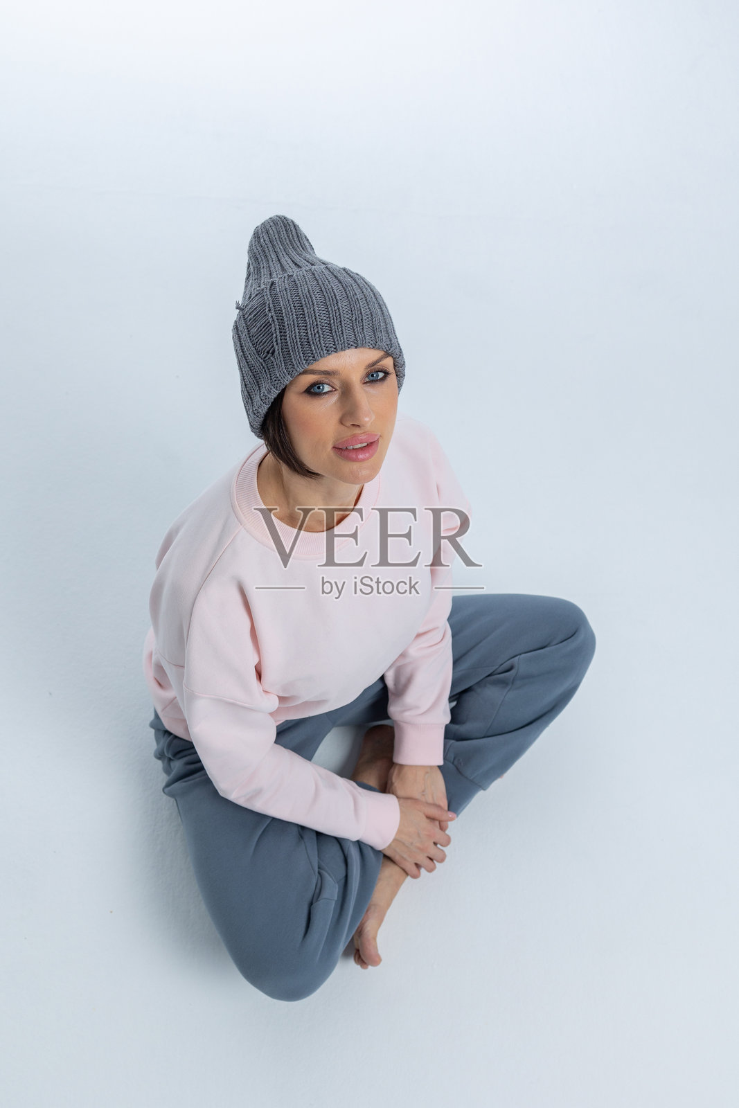 穿着灰色帽子的美丽女性肖像。女孩坐在地板上,双腿交叉。工作室摄影。竖版照片。照片摄影图片