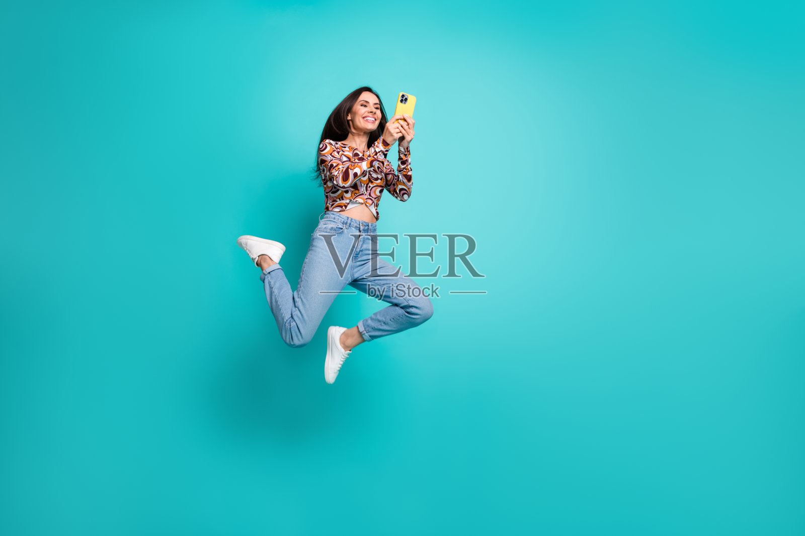 可爱的女孩穿着印花衬衫在空旷的蓝绿色背景上高高跳起,手中拿着聊天设备。照片摄影图片