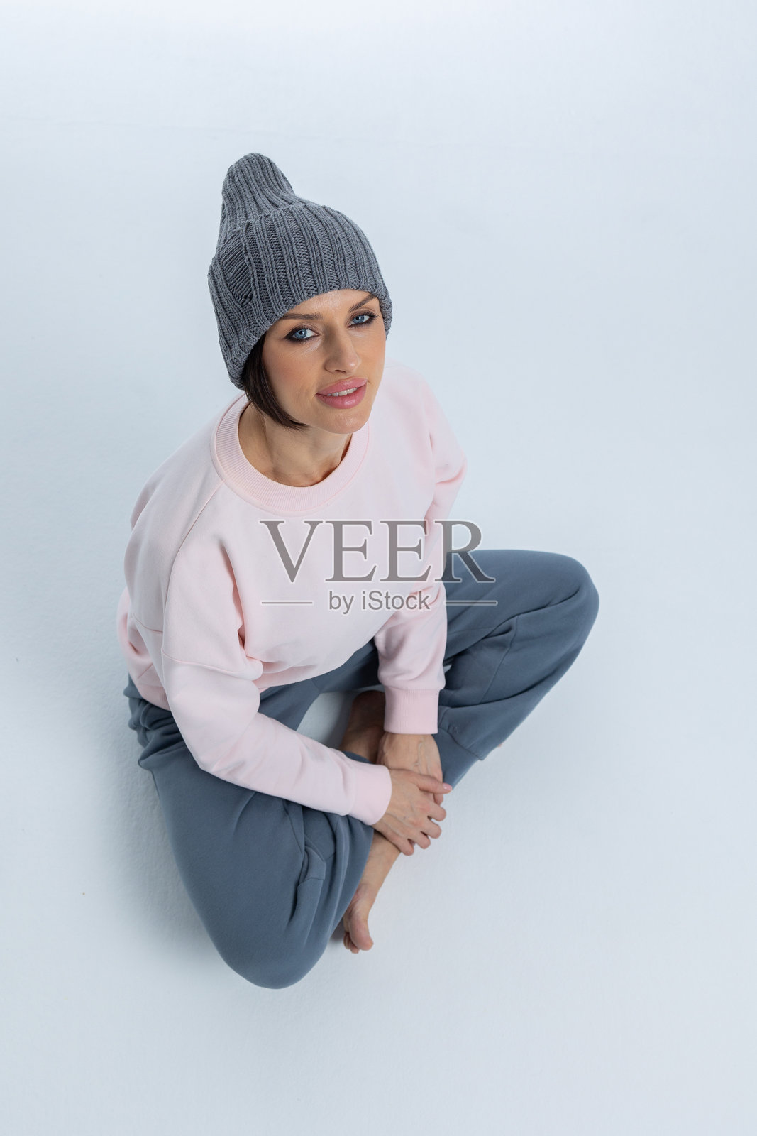 穿着灰色帽子的美丽女性肖像。女孩坐在地板上,双腿交叉。工作室摄影。竖版照片。照片摄影图片