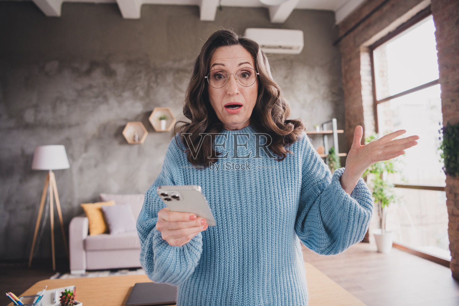 惊讶的老年女性在现代装饰的舒适客厅里看着智能手机,阳光透过窗户洒入室内。照片摄影图片