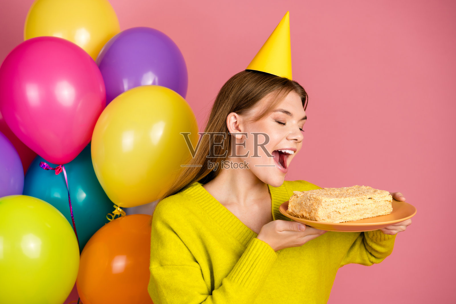 年轻女性穿着黄色毛衣和派对帽,庆祝时与蛋糕和气球在粉色背景下。照片摄影图片