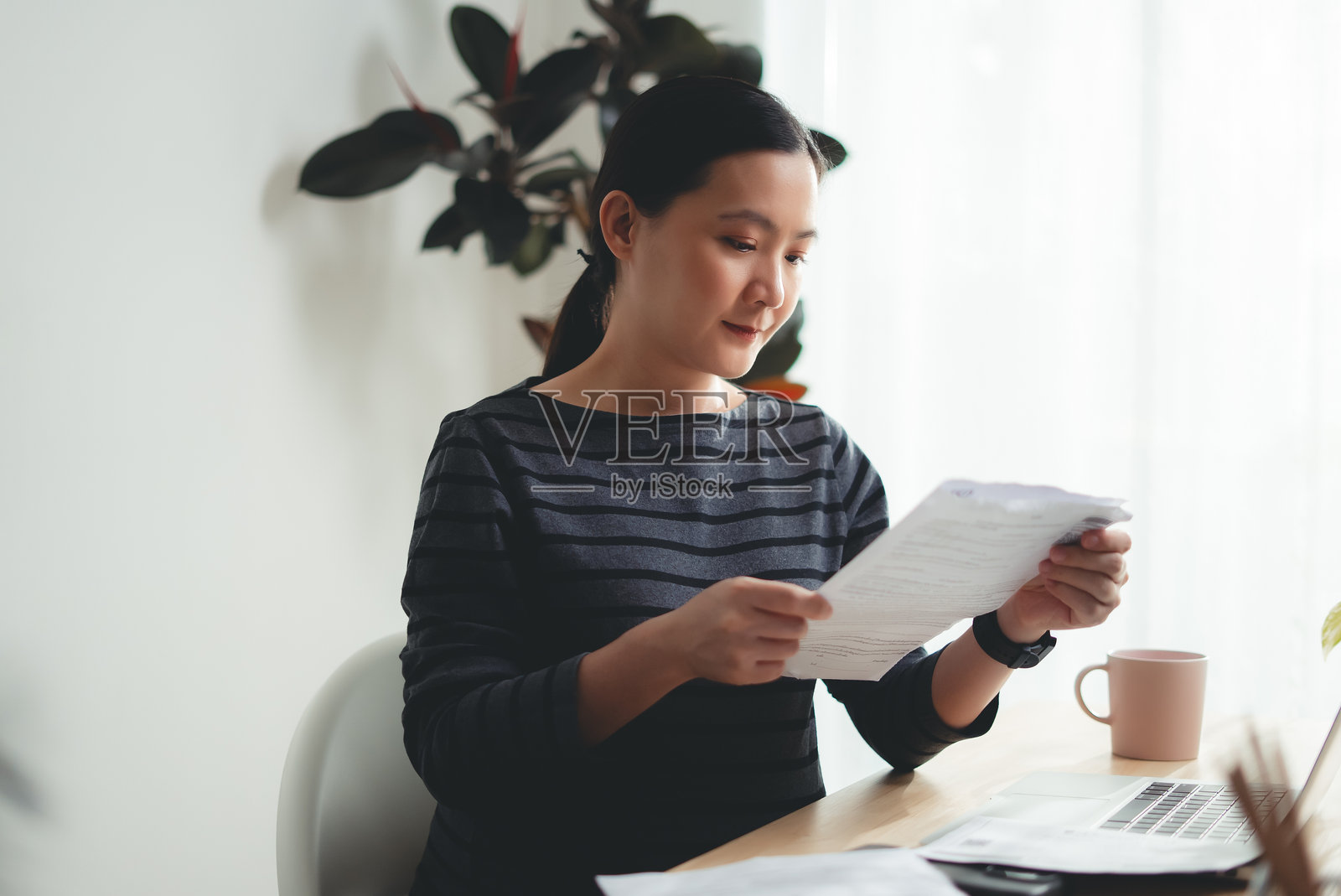 亚洲女性在办公室坐着,手持文件并使用笔记本电脑,享受在线工作的乐趣。照片摄影图片