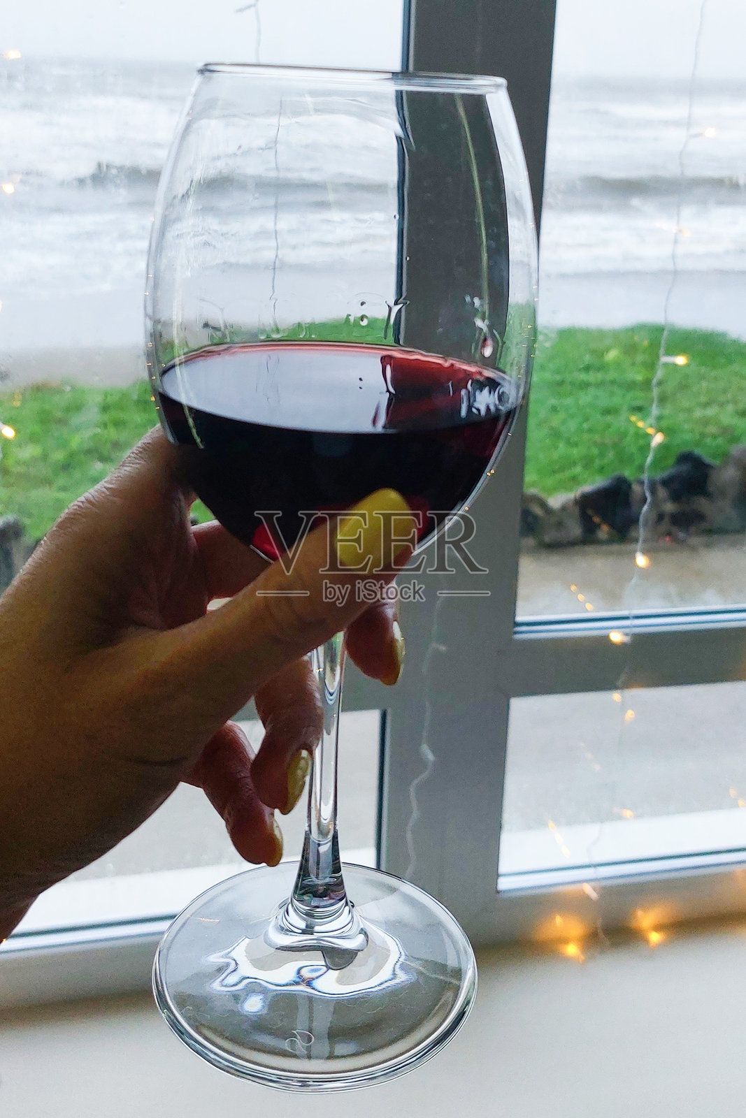 一只涂着黄色指甲油的手握着一杯红酒,窗外是海洋景色和暴风雨天气。照片摄影图片