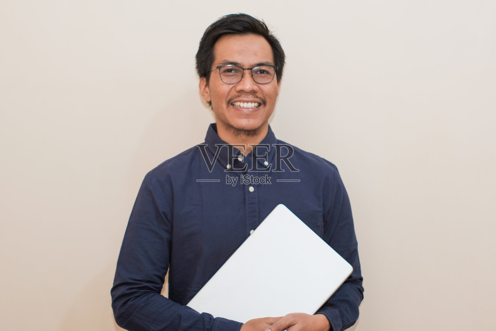 微笑的东南亚男子穿着蓝色衬衫和眼镜,手持笔记本电脑,站在孤立的白色背景前。照片摄影图片