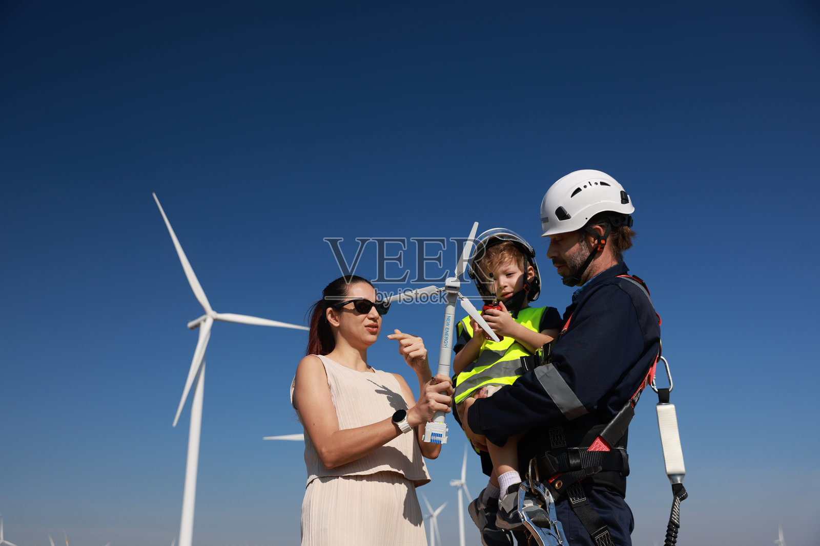 阳光明媚的日子里,父亲工程师无忧无虑地牵着儿子的手,与妻子一起在风车田野中。可再生能源、热爱自然、家庭和电力的概念。照片摄影图片