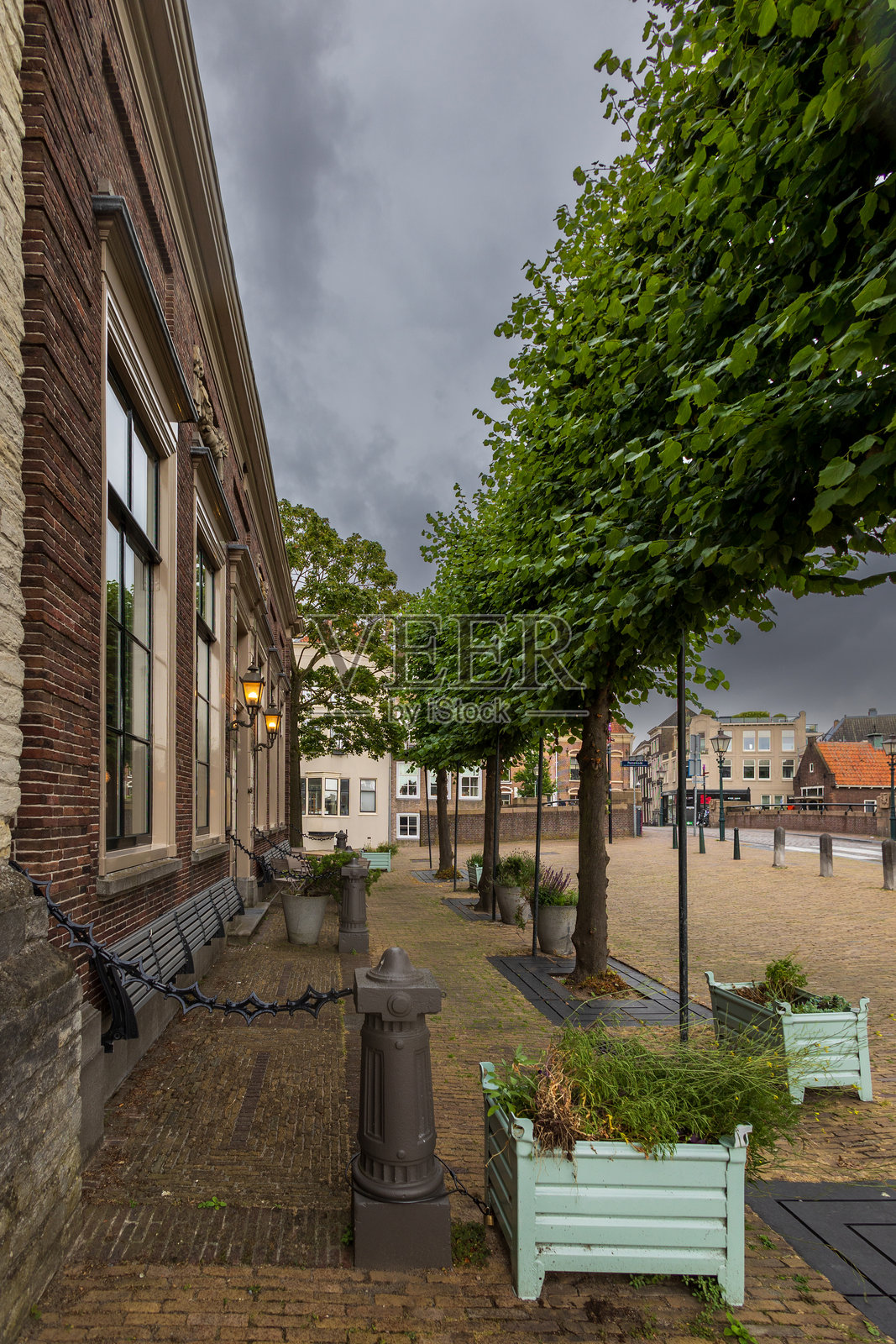 一条安静的鹅卵石街道,绿树成荫,长椅沿着一座砖砌建筑的侧面,天空阴云密布。复古风格的灯笼为这幅历史悠久的城市景观增添了魅力。多德雷赫特风景。照片摄影图片