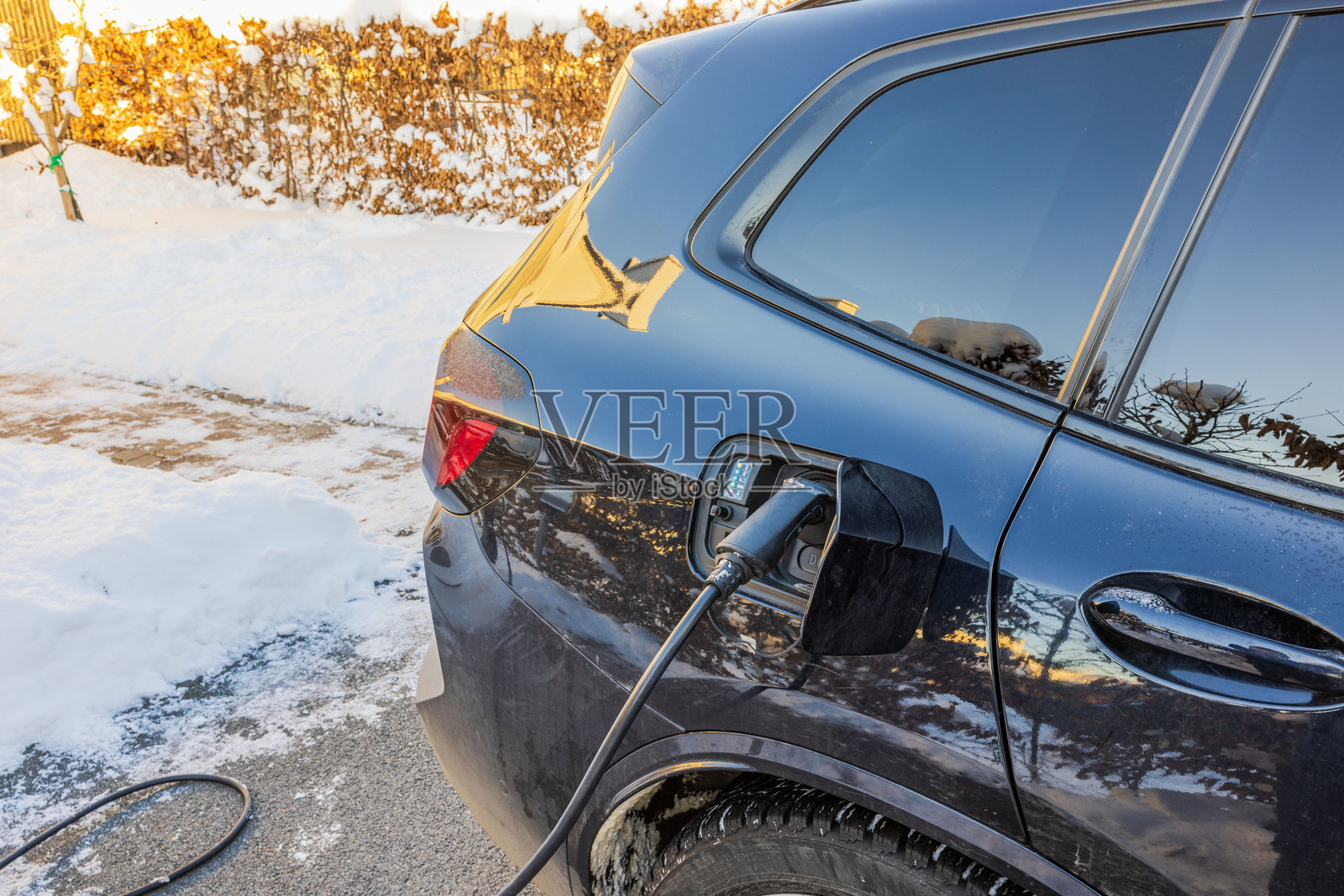 冬季雪地车道上的电动车充电,充电线连接到后部充电口。瑞典。照片摄影图片