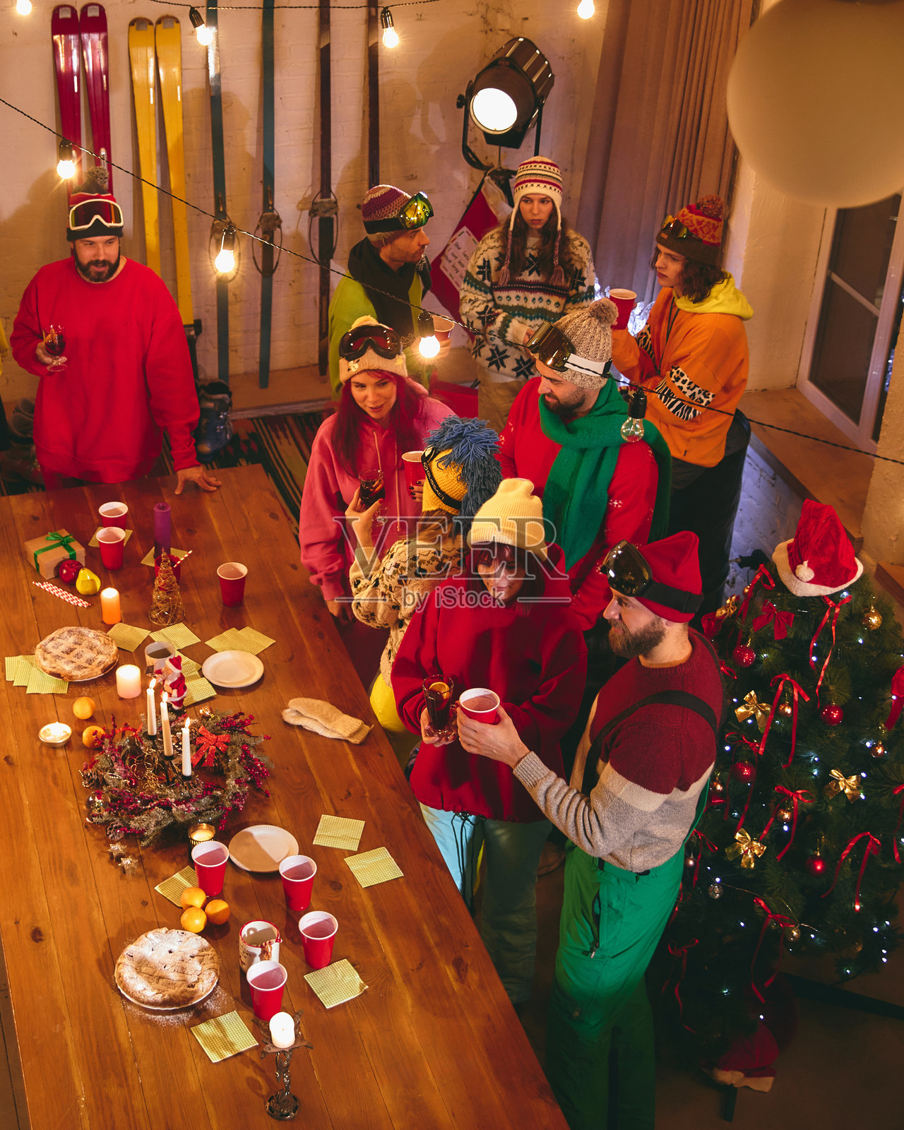 温暖的节日气氛充满房间,朋友们穿着鲜艳的滑雪装备,在装饰精美的桌子旁举杯庆祝,享用小吃,桌上有节日灯饰、蜡烛和糕点。照片摄影图片