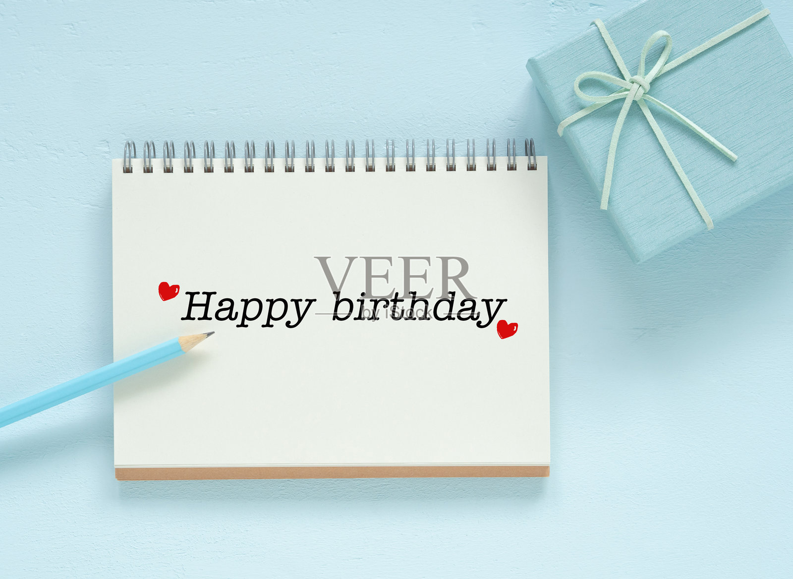 从上方俯视,空白笔记本页面上写着“生日快乐”的文字,旁边有铅笔和礼品盒,背景为蓝色木板。留有空间供您填写文字。为写生日祝福的创意构思。照片摄影图片